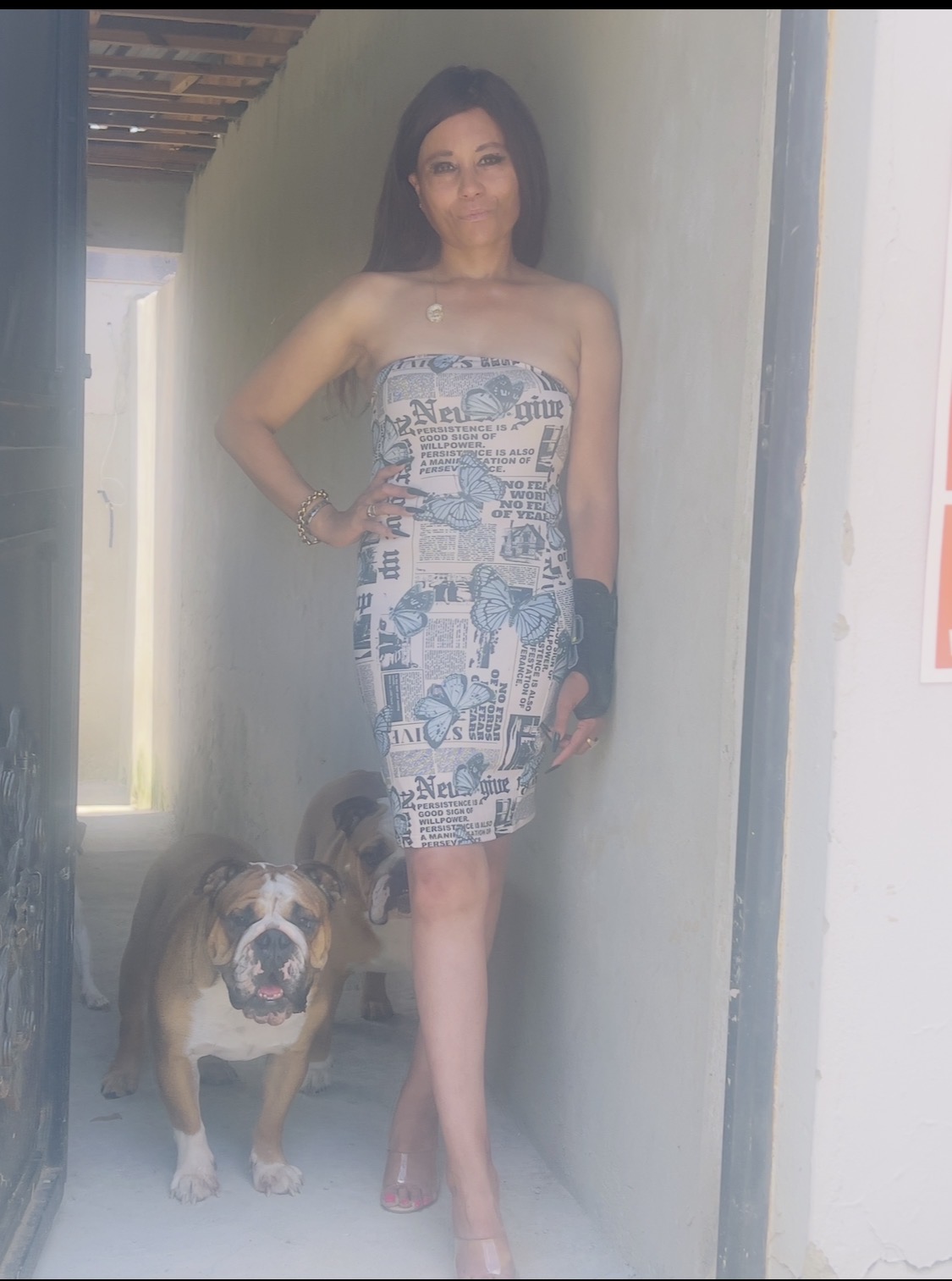 Cass in a strapless newspaper-print mini dress with pale pink heels, posing in a doorway with Stretford the bulldog