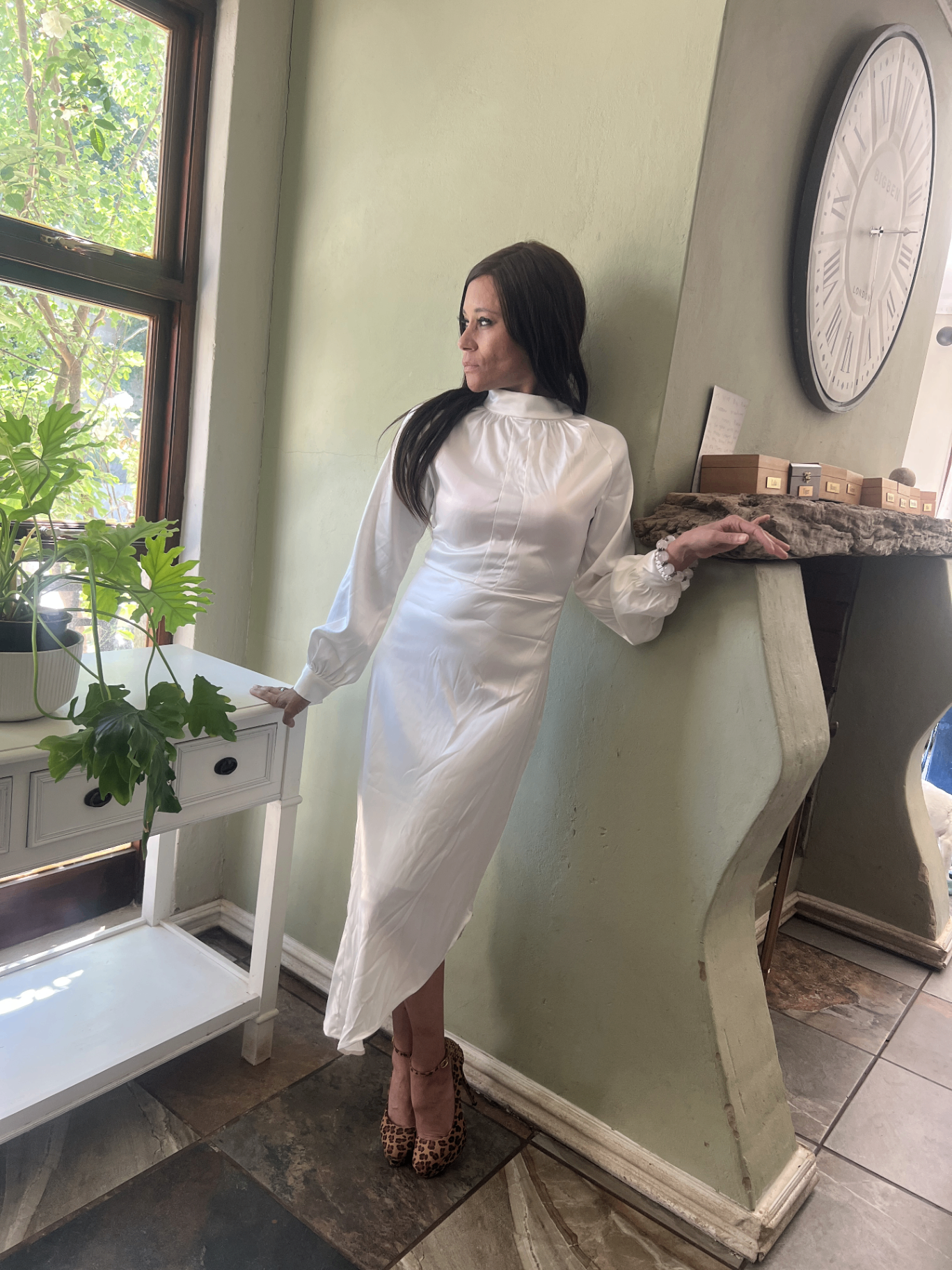Cass in a white satin midi dress and leopard-print heels, leaning against a sage-green wall in a sunlit Jo'burg room.