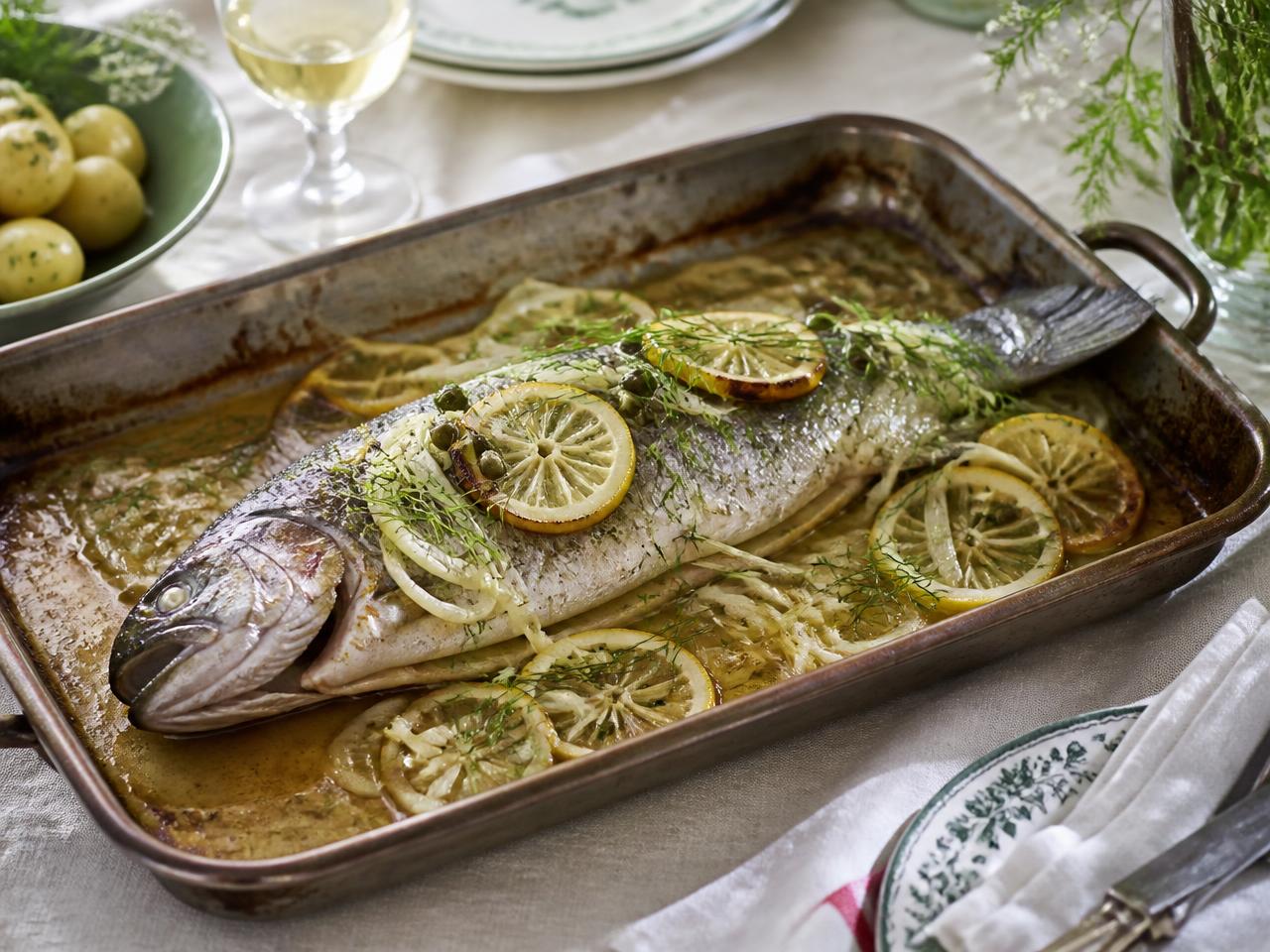 Whole baked trout on a roasting tray with shaved fennel, capers, lemon slices and dill in buttery pan juices