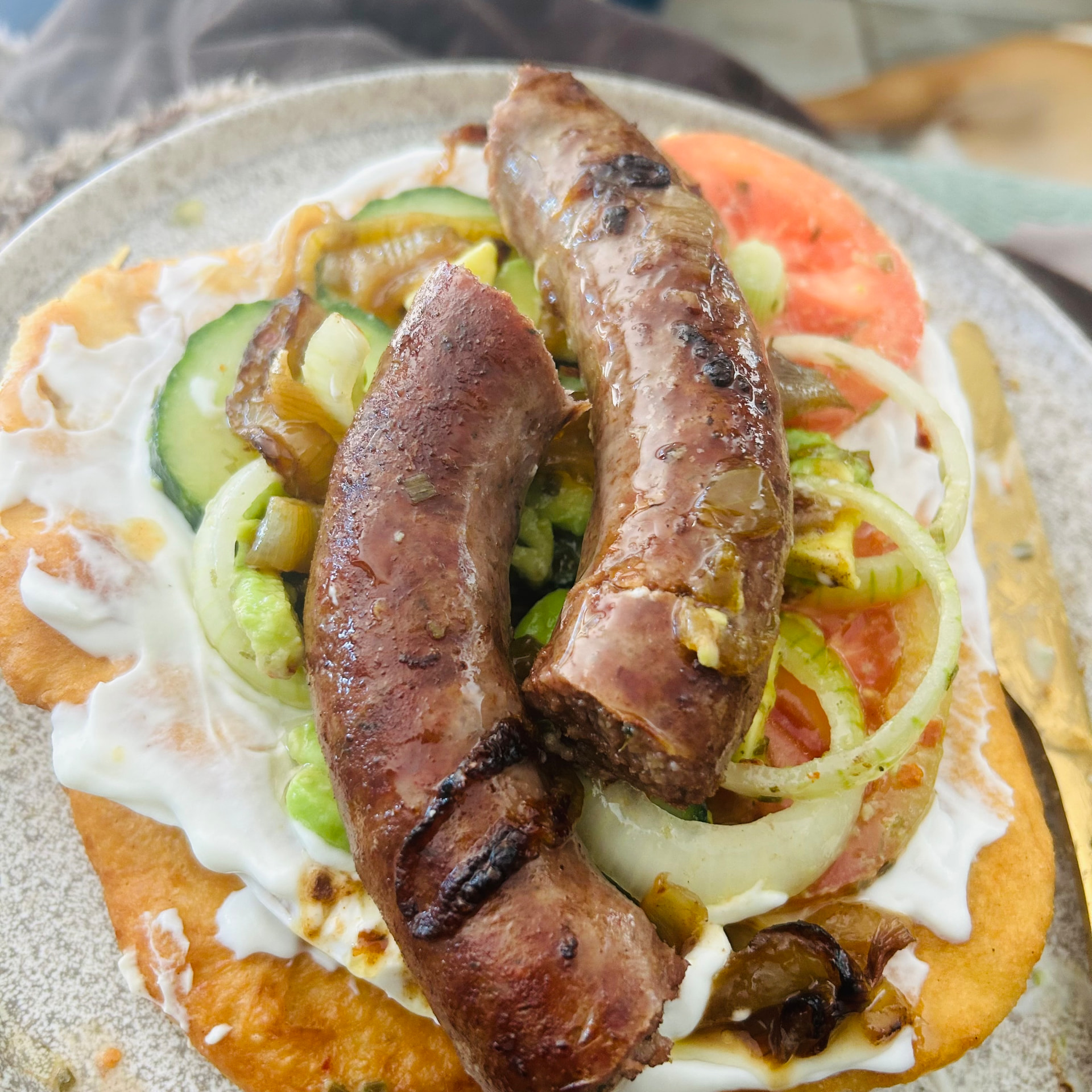 Two plump browned boerewors sausages resting on lángos with labneh, avocado, cucumber and tomato salad