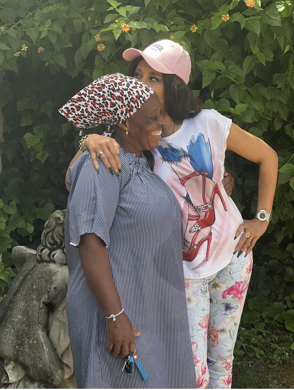 Cass laughing, arm wrapped around Ntombi who is hugging her close — a tender, sunlit garden moment between the two of them