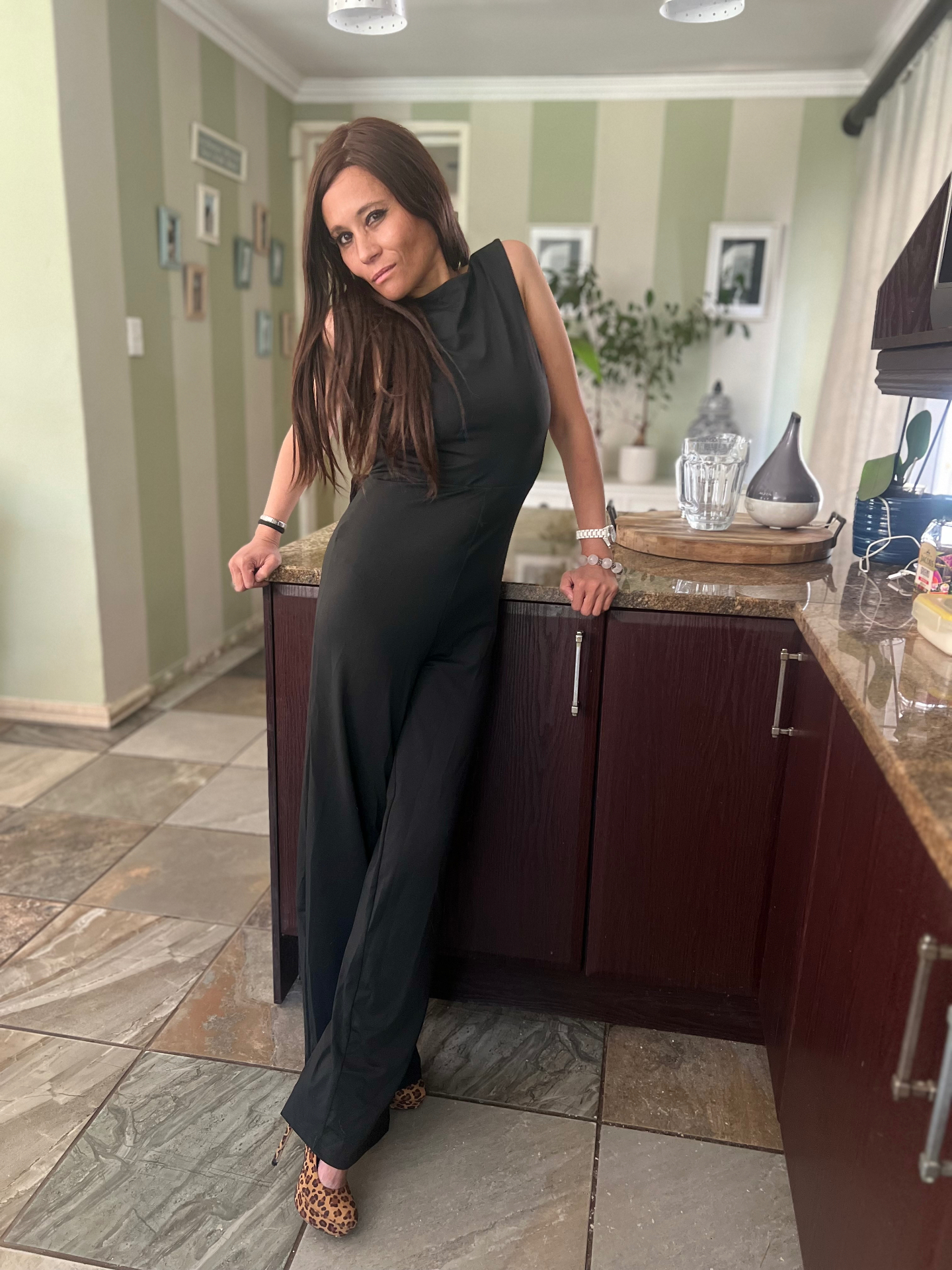 Cass in a sleeveless high-neck black jumpsuit with leopard-print heels, leaning against a kitchen counter