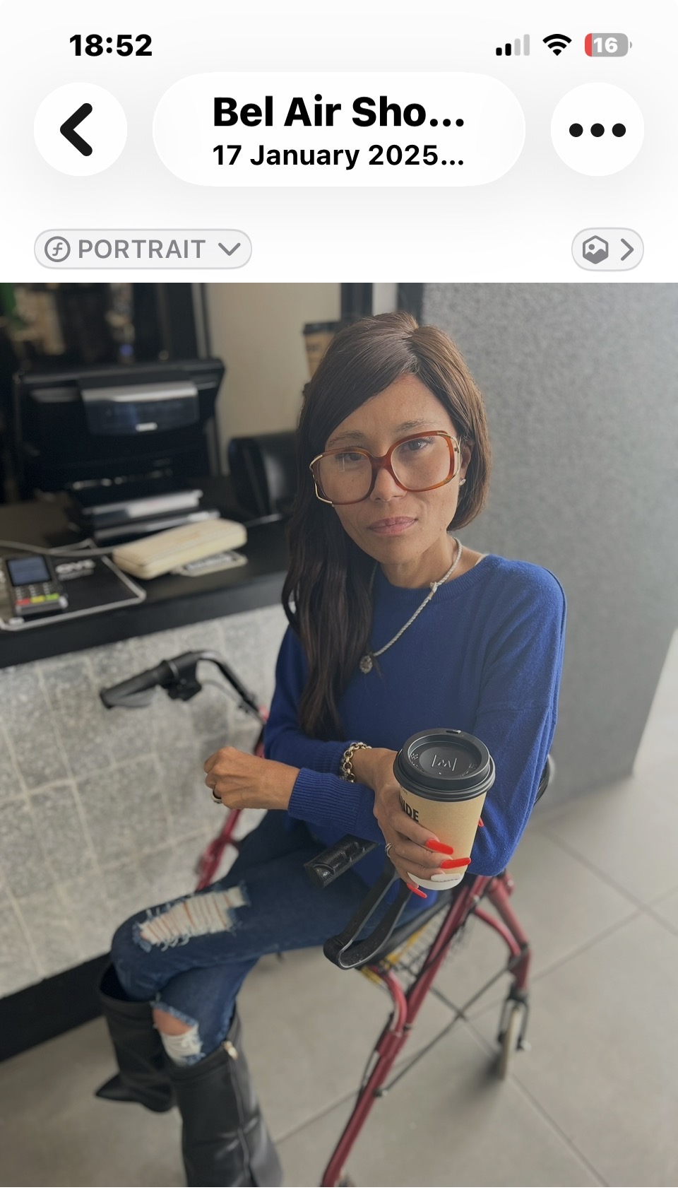 Cass in oversized tortoiseshell glasses and a cobalt-blue jumper, sitting on a red walker with a grande cappuccino in hand, at a Bel Air showroom