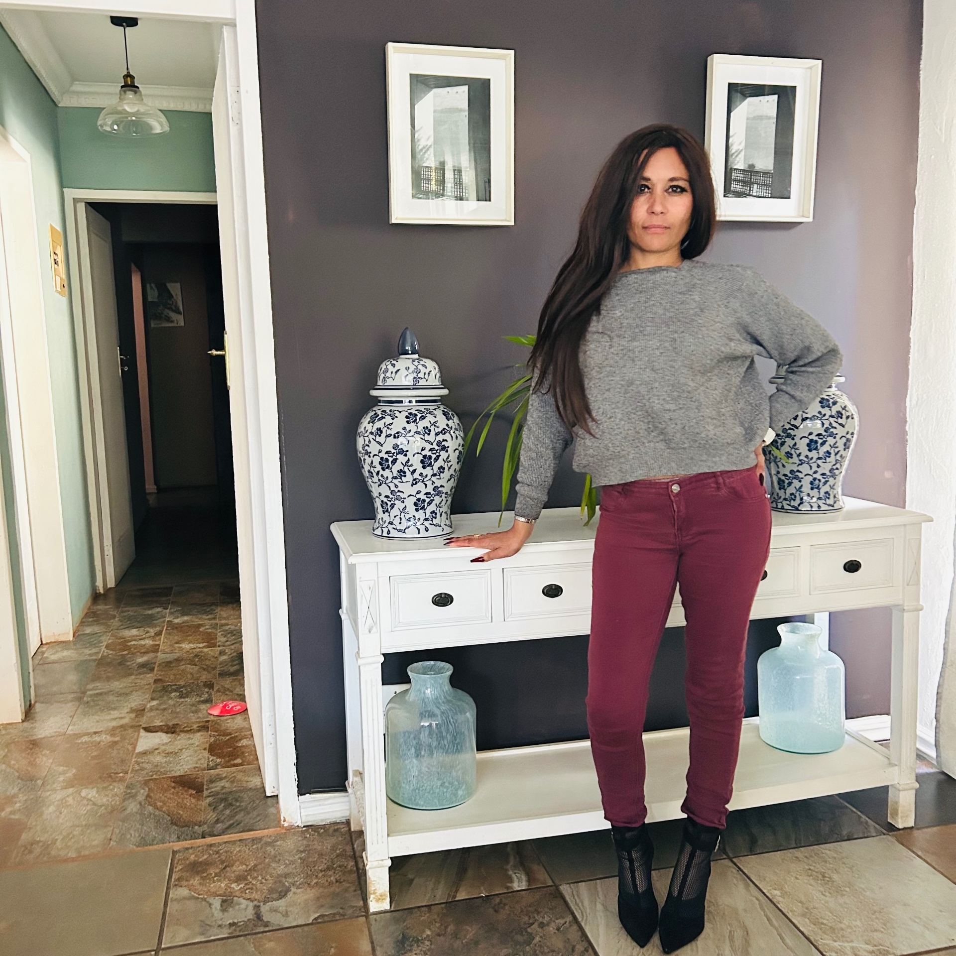 Cass in a grey cropped knit, burgundy skinny jeans and black pencil-heel mesh ankle boots, posing next to a white console with blue-and-white ginger jars