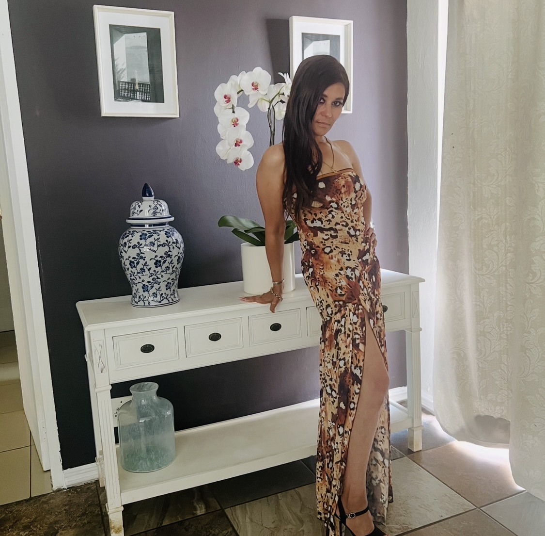 Cass leaning on a white console table in the leopard-print maxi dress, with orchids and a blue-and-white ginger jar