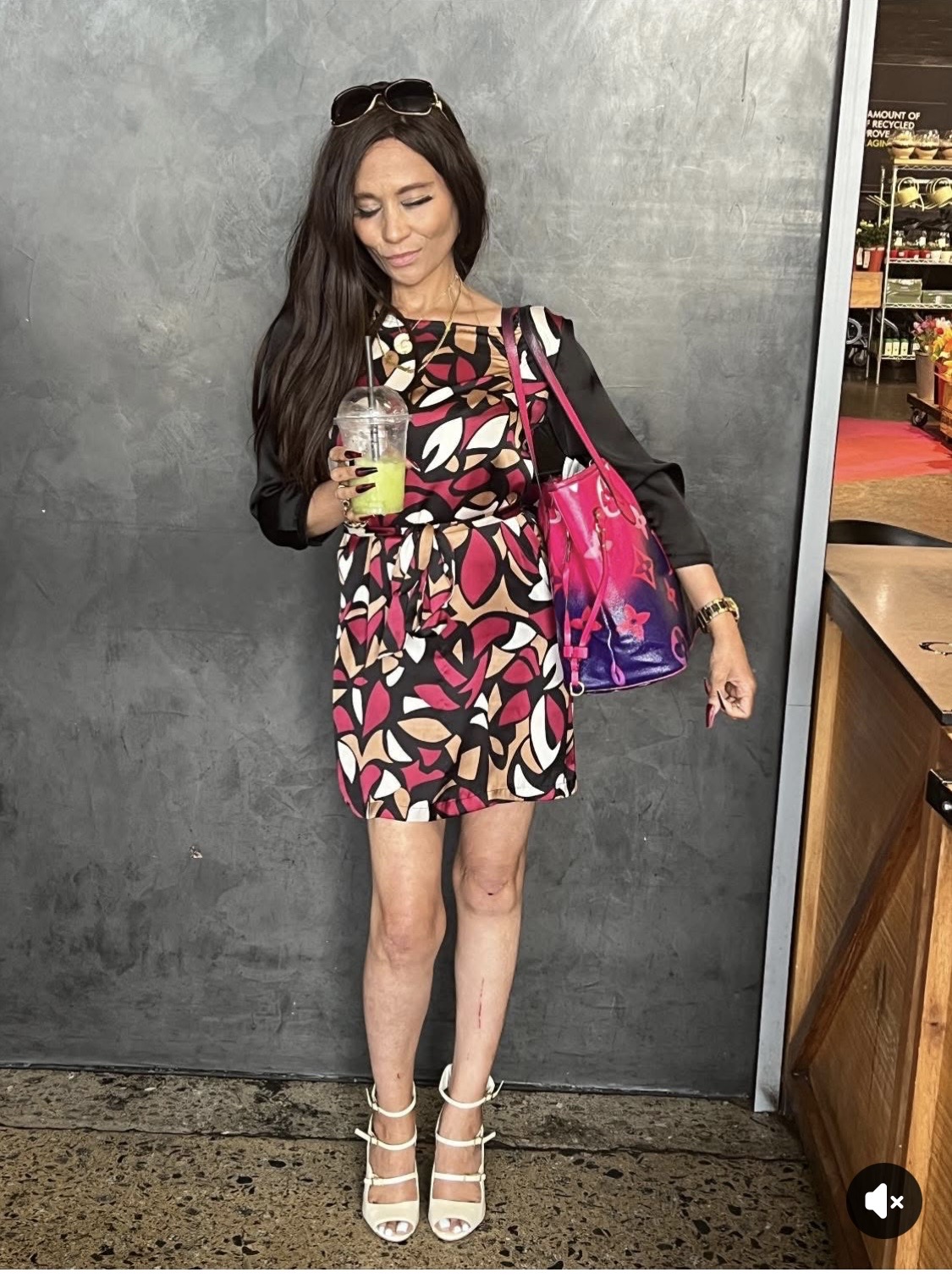 Cass in a bold pink, black, tan and white abstract-print mini dress, massive Louis Vuitton tote, and white strappy Socrati heels, holding a lemon-and-mint crushed ice-tea