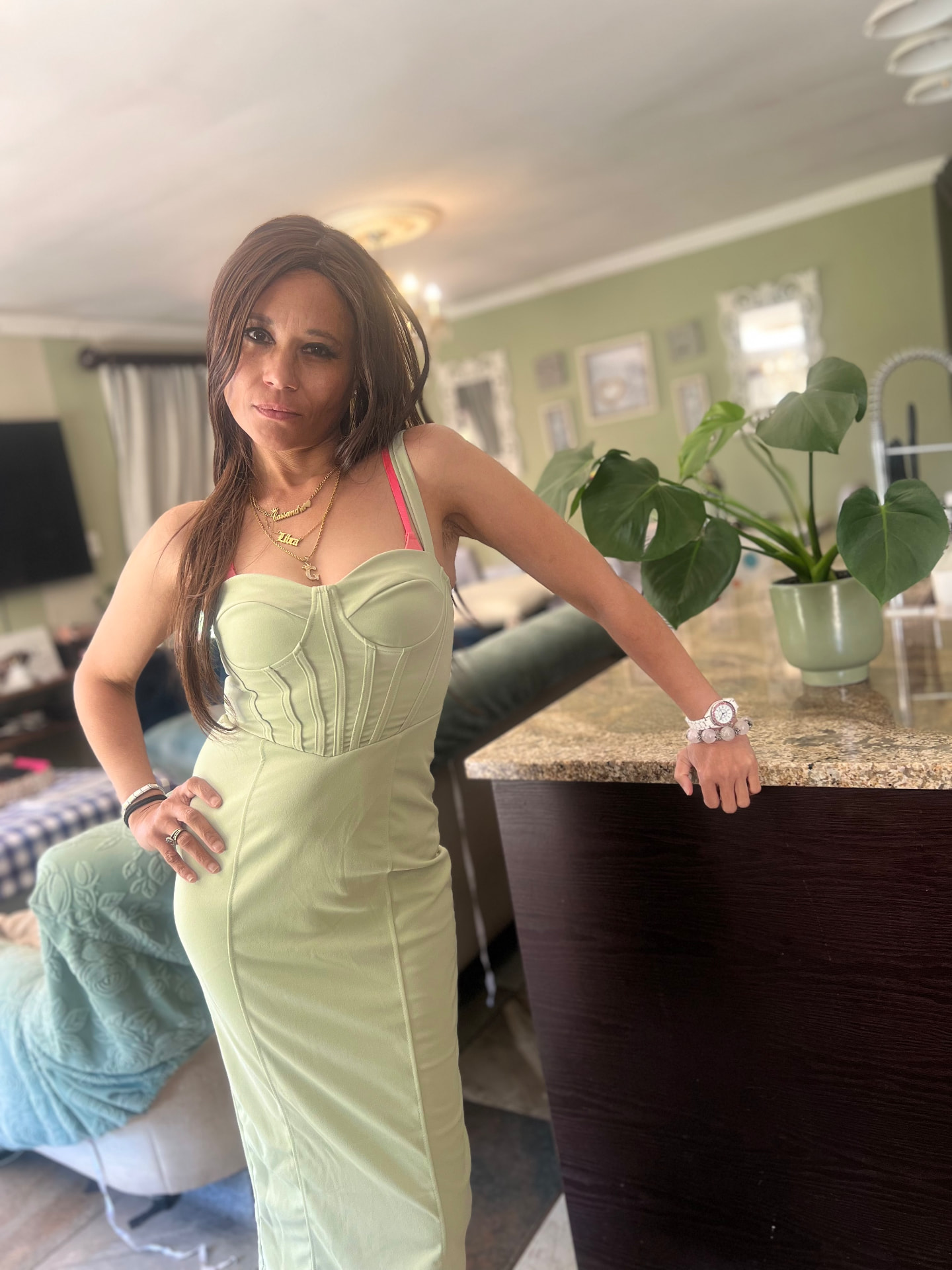Cass in a powder-mint corset midi dress leaning on the kitchen island beside a monstera
