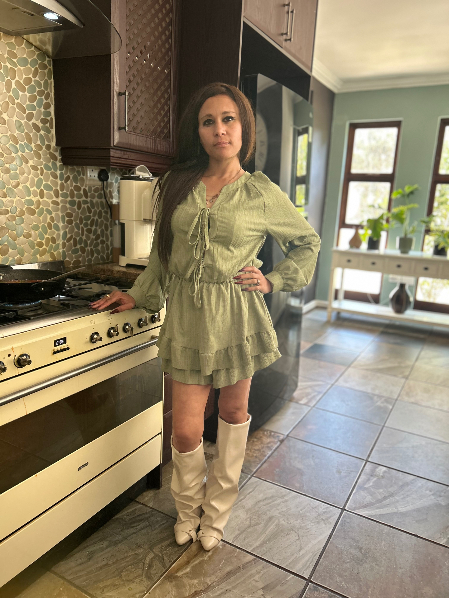 Cass in an olive-green tie-front ruffle playsuit and cream cone-heel knee boots, standing in her kitchen