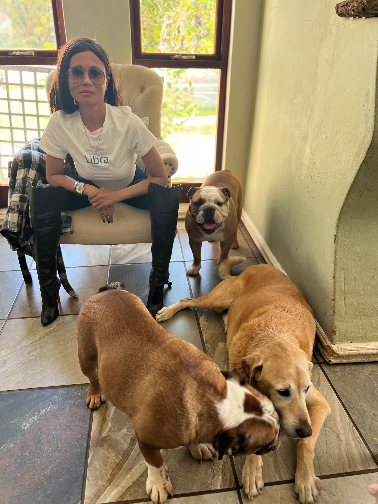 Cass in a white Libra tee, denim and black over-the-knee leather boots, seated in a wing chair with four dogs around her