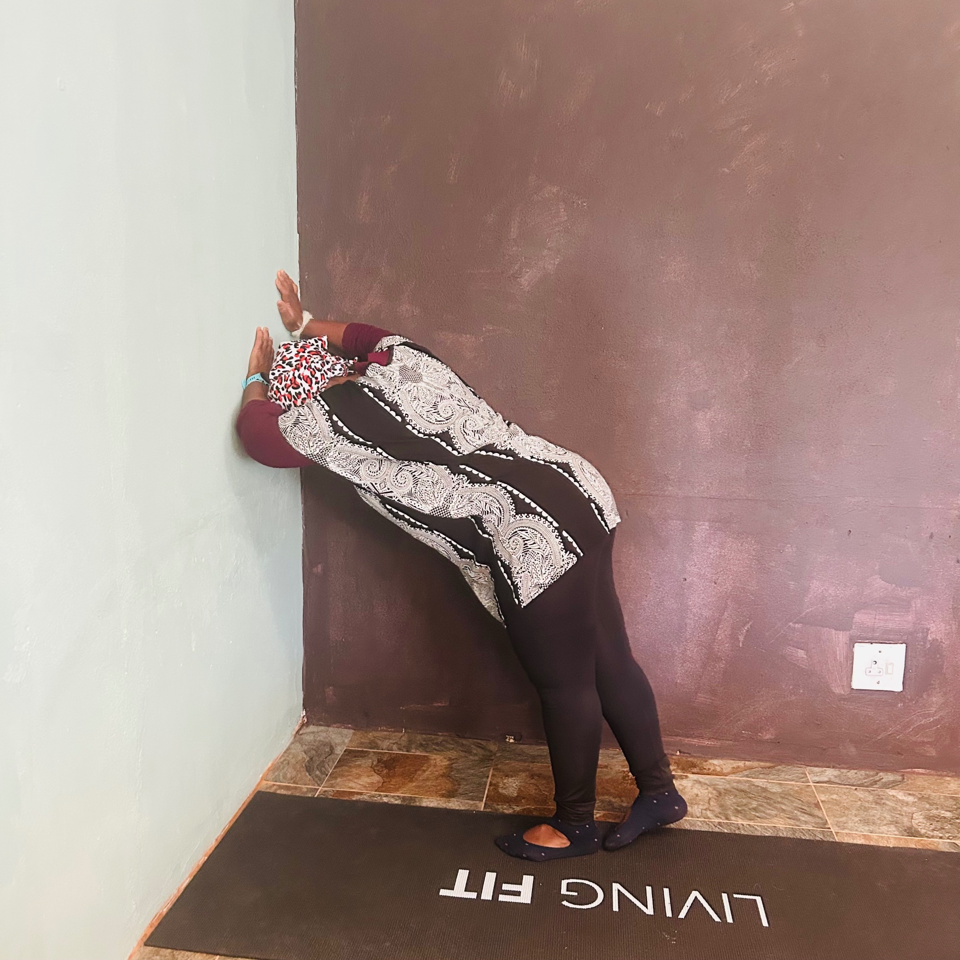 Ntombi bent forward against a burgundy wall, palms flat, mid tai chi stretch, on a Living Fit mat.