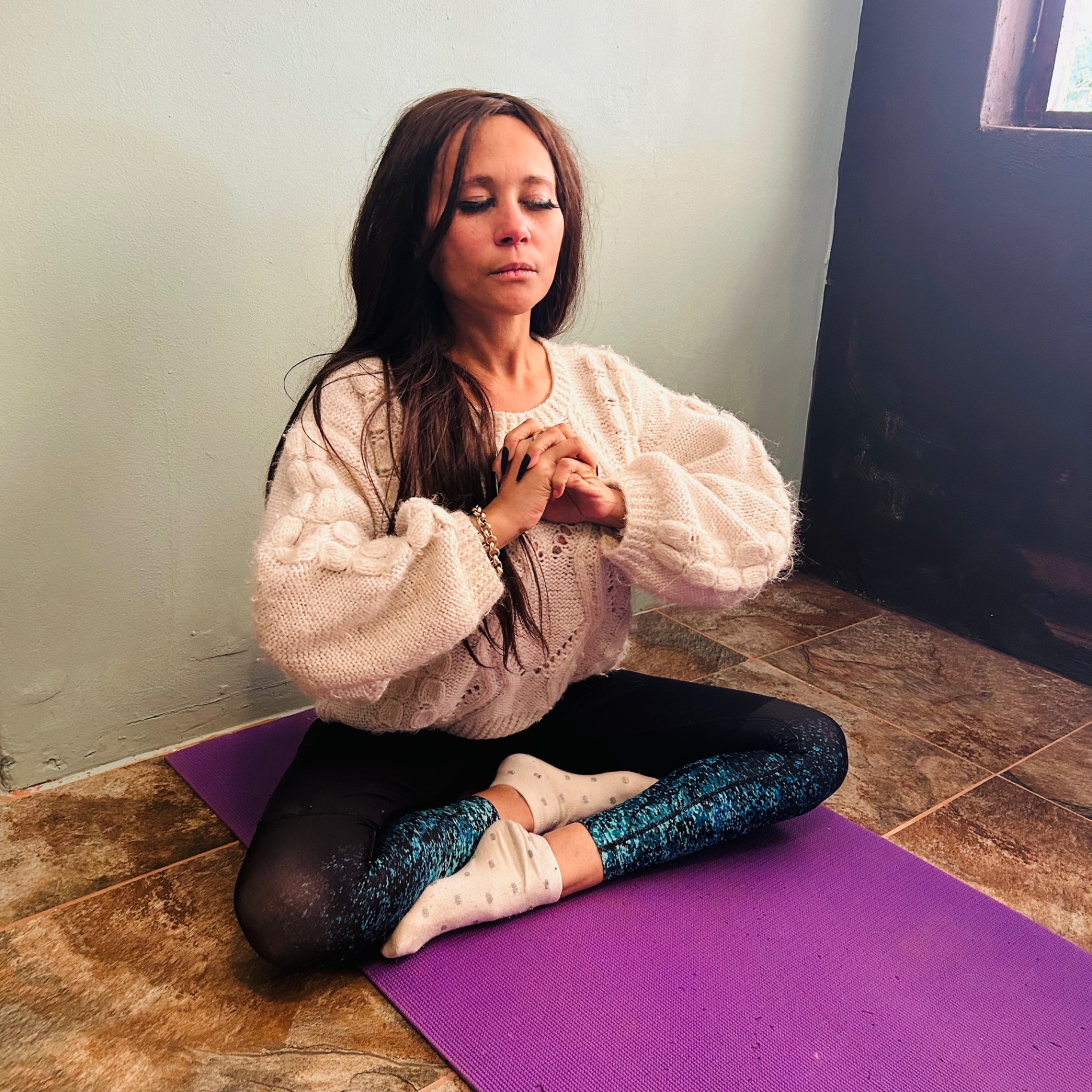 Cass sitting cross-legged in meditation, hands at heart centre, on a purple yoga mat