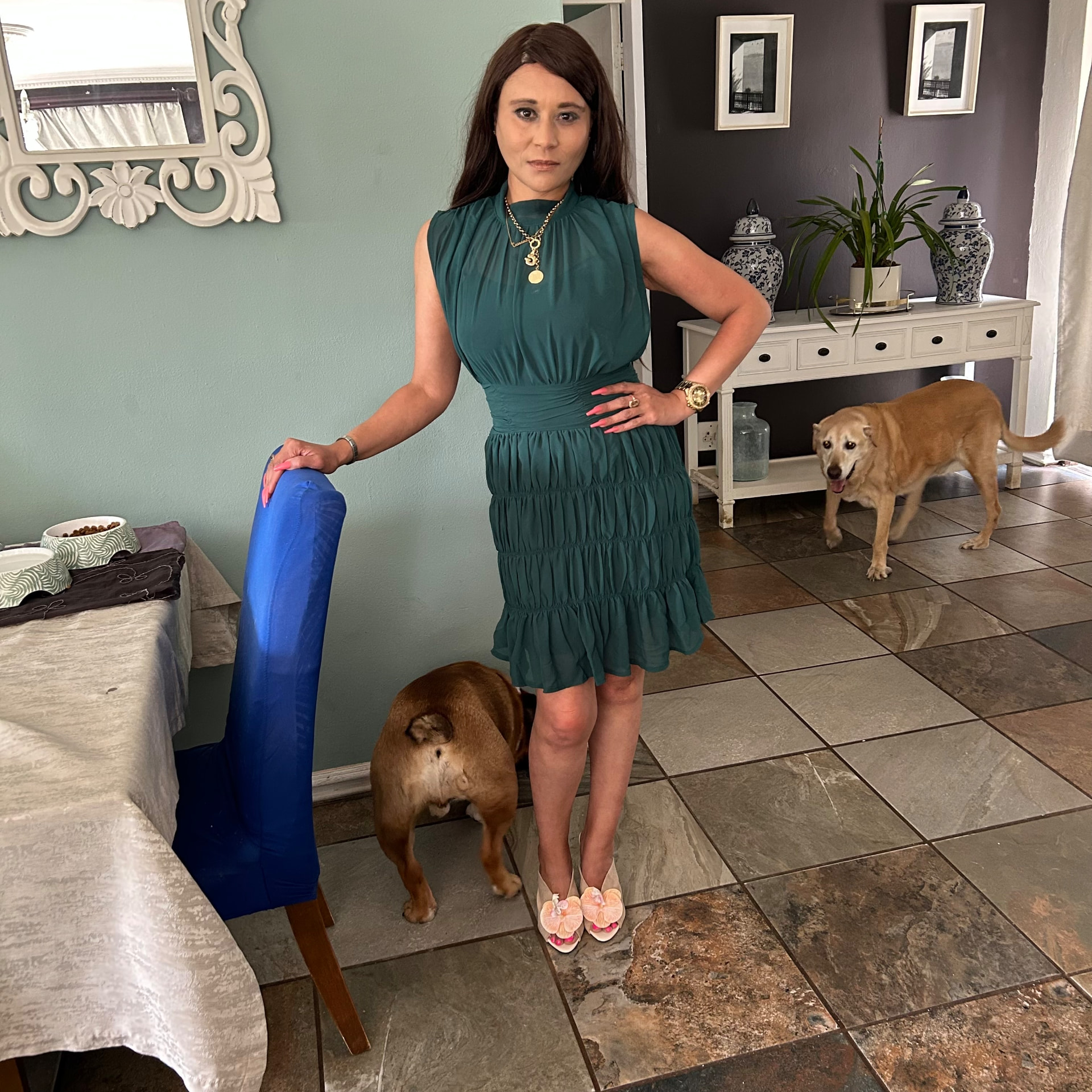 Cass in an emerald green ruched sleeveless dress with shell-flower heels, one hand on a blue chair, with Stretford at her feet