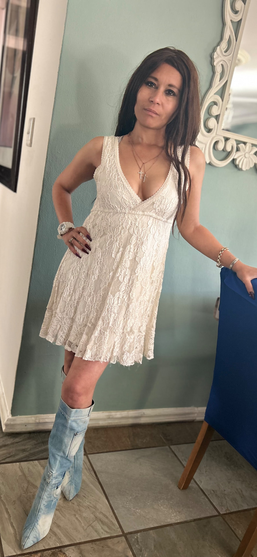 Cass in an ivory lace V-neck mini dress and light-wash denim knee boots, standing in a room with sage walls and an ornate white mirror