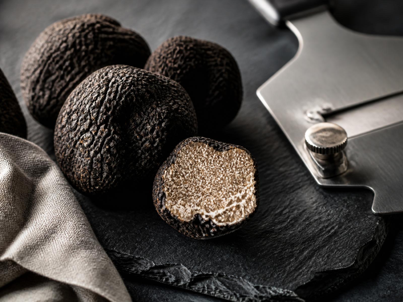Whole black winter truffles on a slate board with a truffle shaver, one halved to show its marbled interior