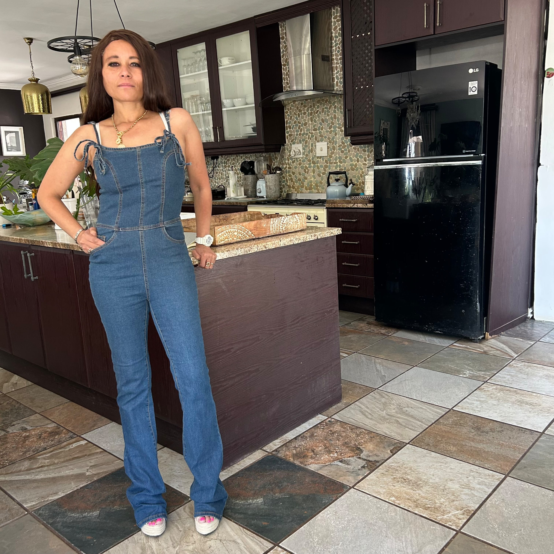 Cass in the denim jumpsuit with tie straps, one hand on her hip in the kitchen