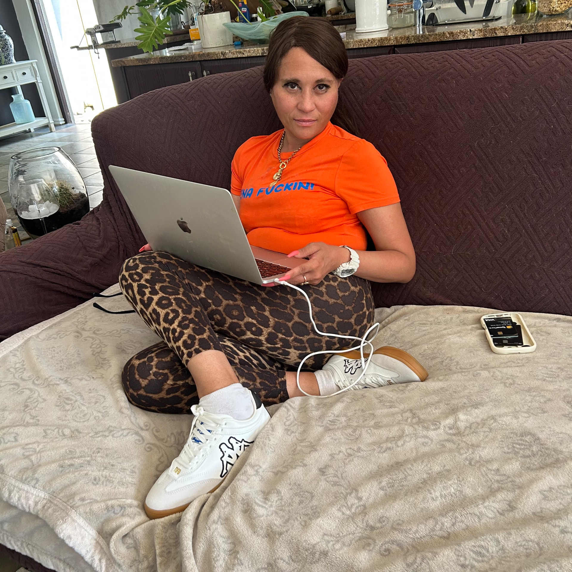 Cass on the couch with a MacBook on her lap, wearing an orange tee with blue lettering, leopard leggings and white Kappa sneakers