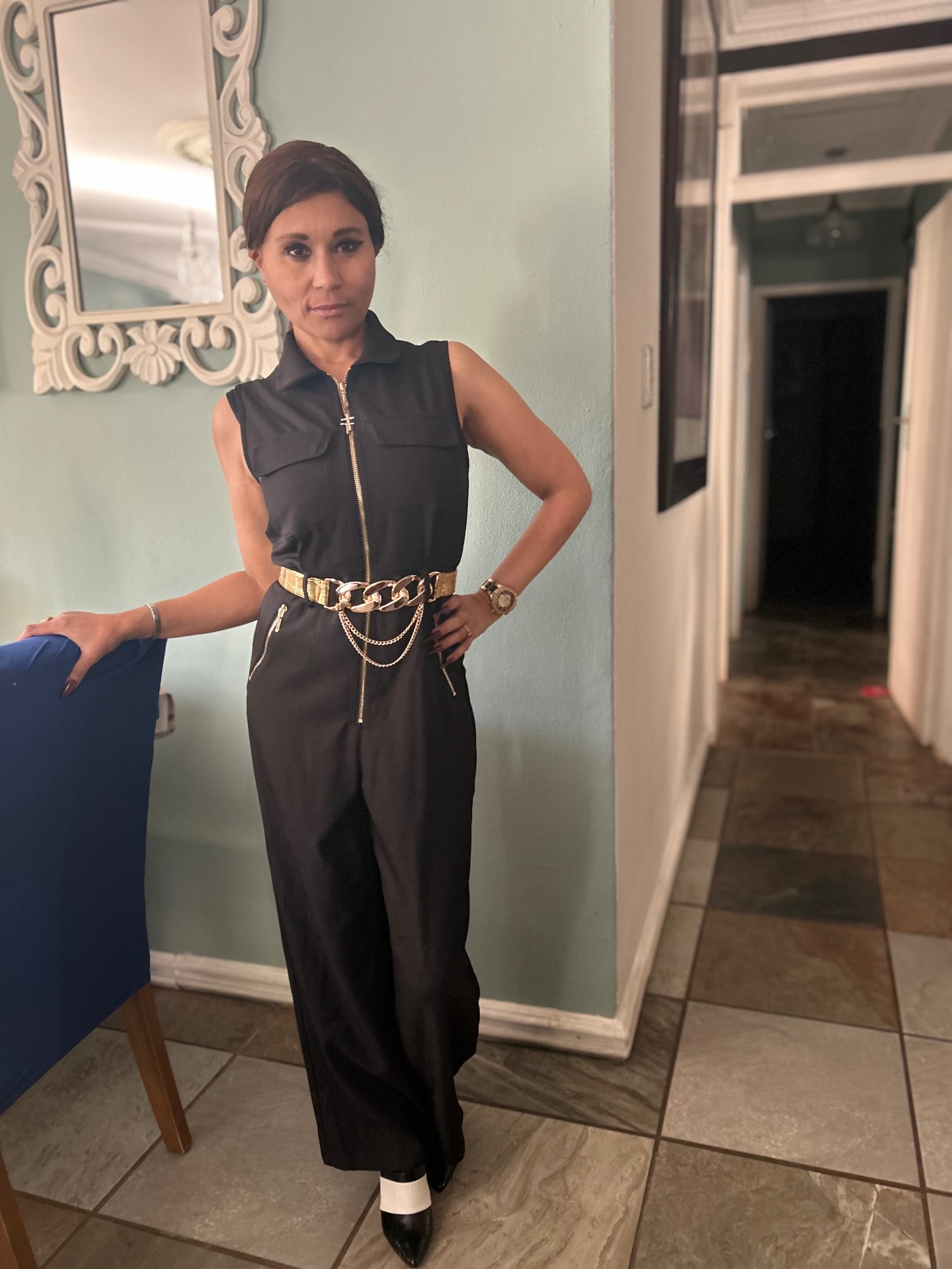 Full-length front view of Cass in the black jumpsuit with hand on hip, gold chain belt catching the light