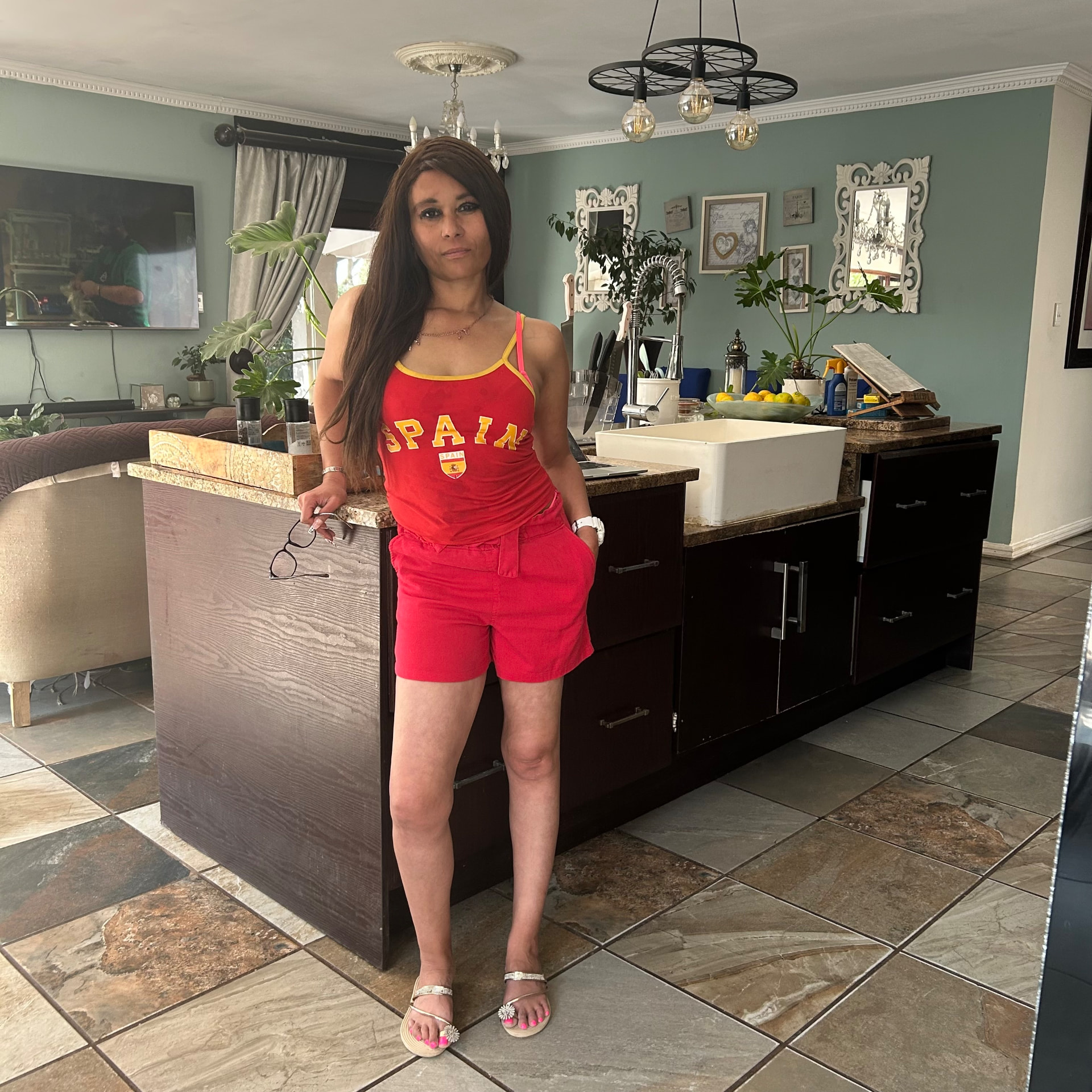 Cass in the red Spain outfit, standing full length in the kitchen with sage walls and pendant lights