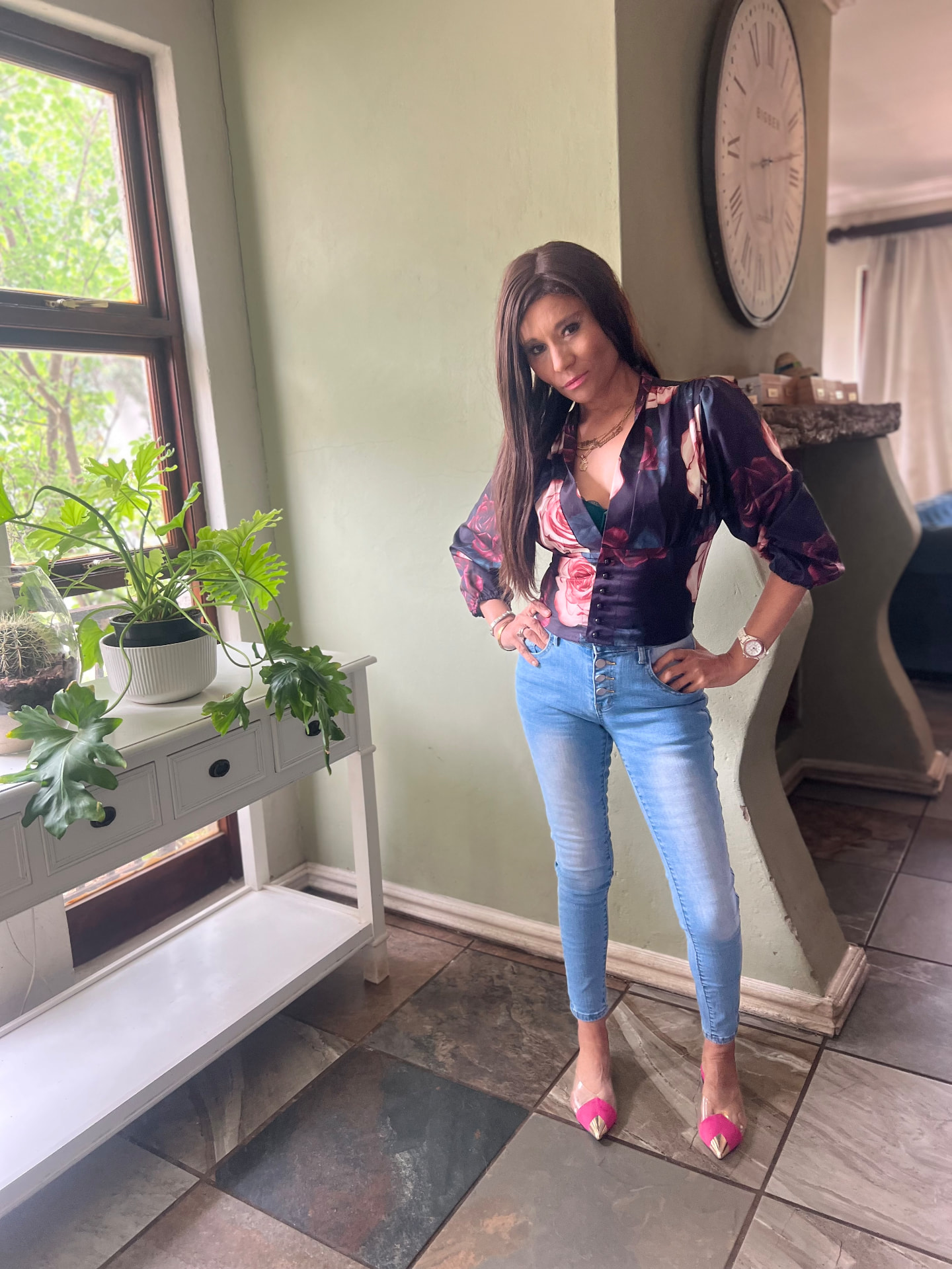 Cass in a dark floral rose blouse with light blue skinny jeans and pink pointed flats, beside a green wall and a large clock