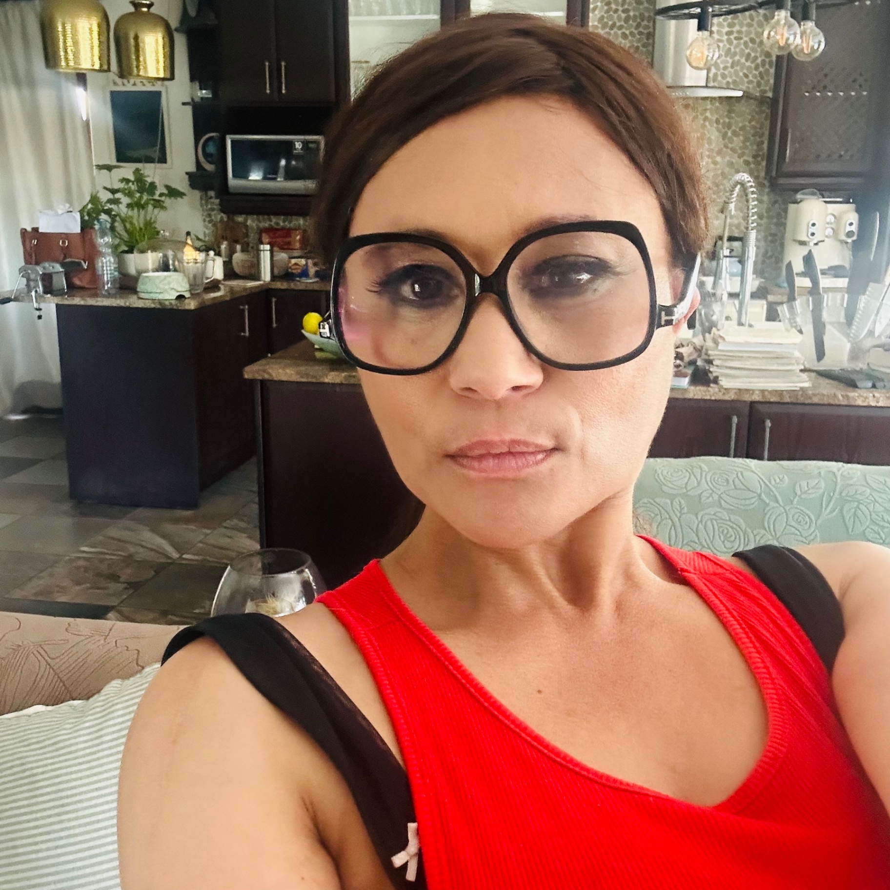 Cass in oversized black square Tom Ford-style sunglasses and red vest, kitchen behind her