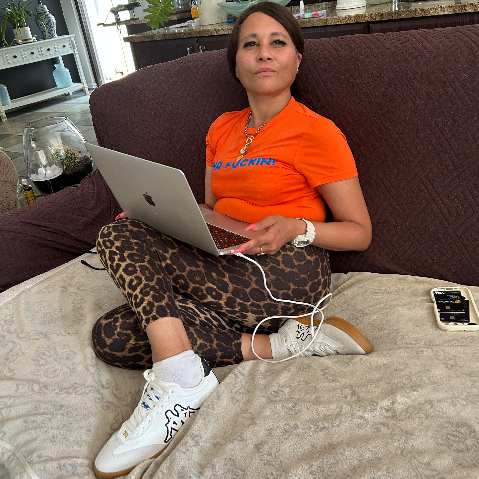 Cass looking at the camera from the couch in the orange tee, leopard leggings and Kappa sneakers