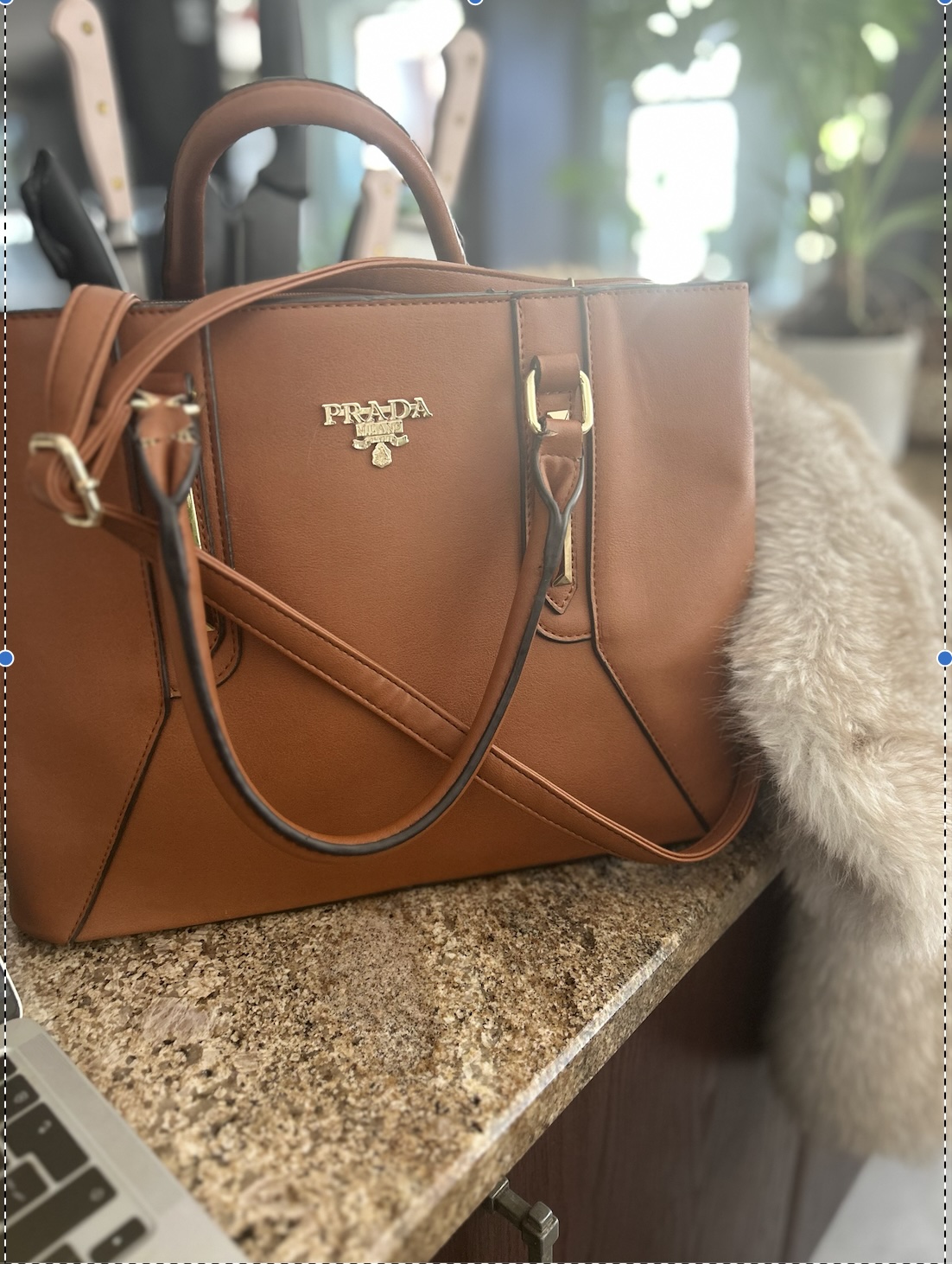 Tan leather Prada tote with gold hardware on a granite counter, faux fur throw beside it