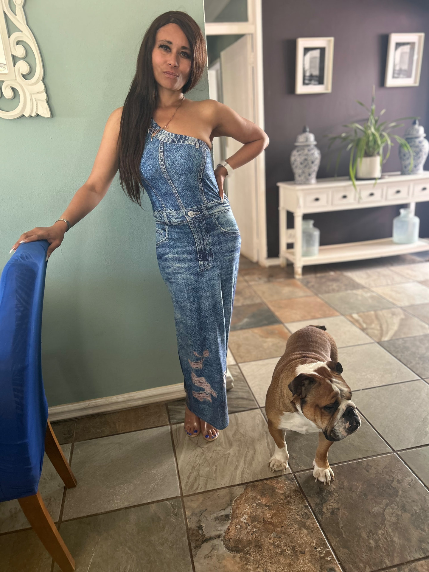 Cass in a one-shoulder acid-wash denim maxi jumpsuit with clear strappy heels against a sage green wall