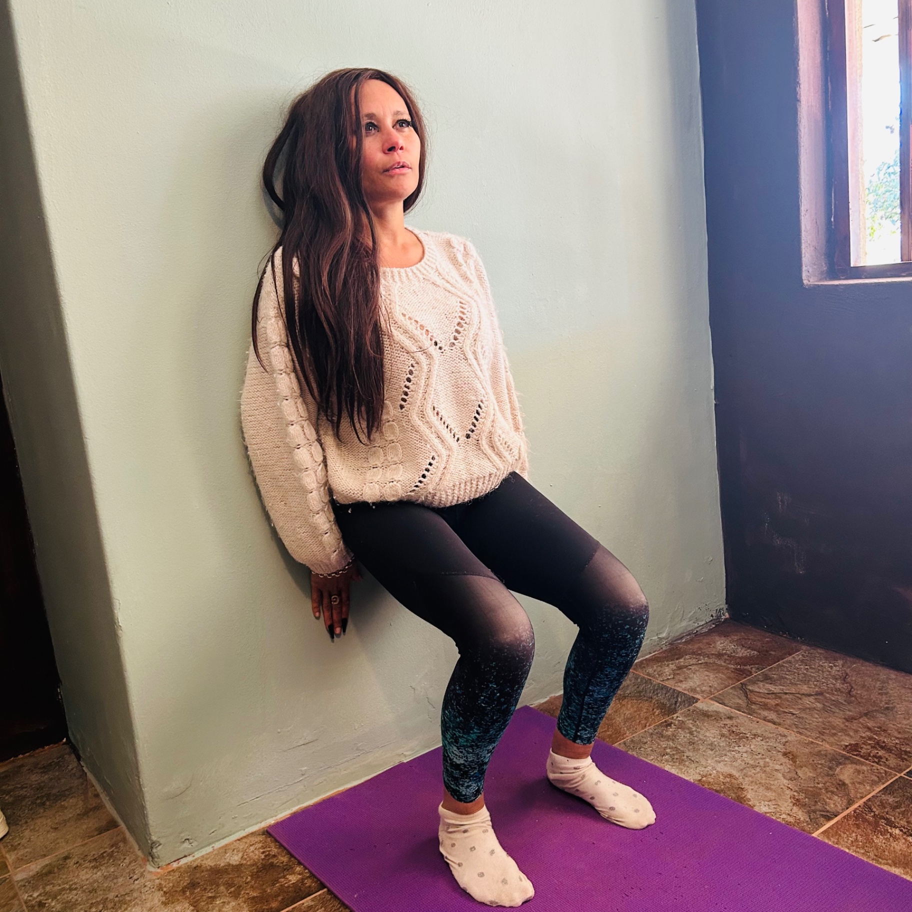 Cass in a cream cable-knit jumper and blue printed leggings, doing a wall sit on a purple yoga mat