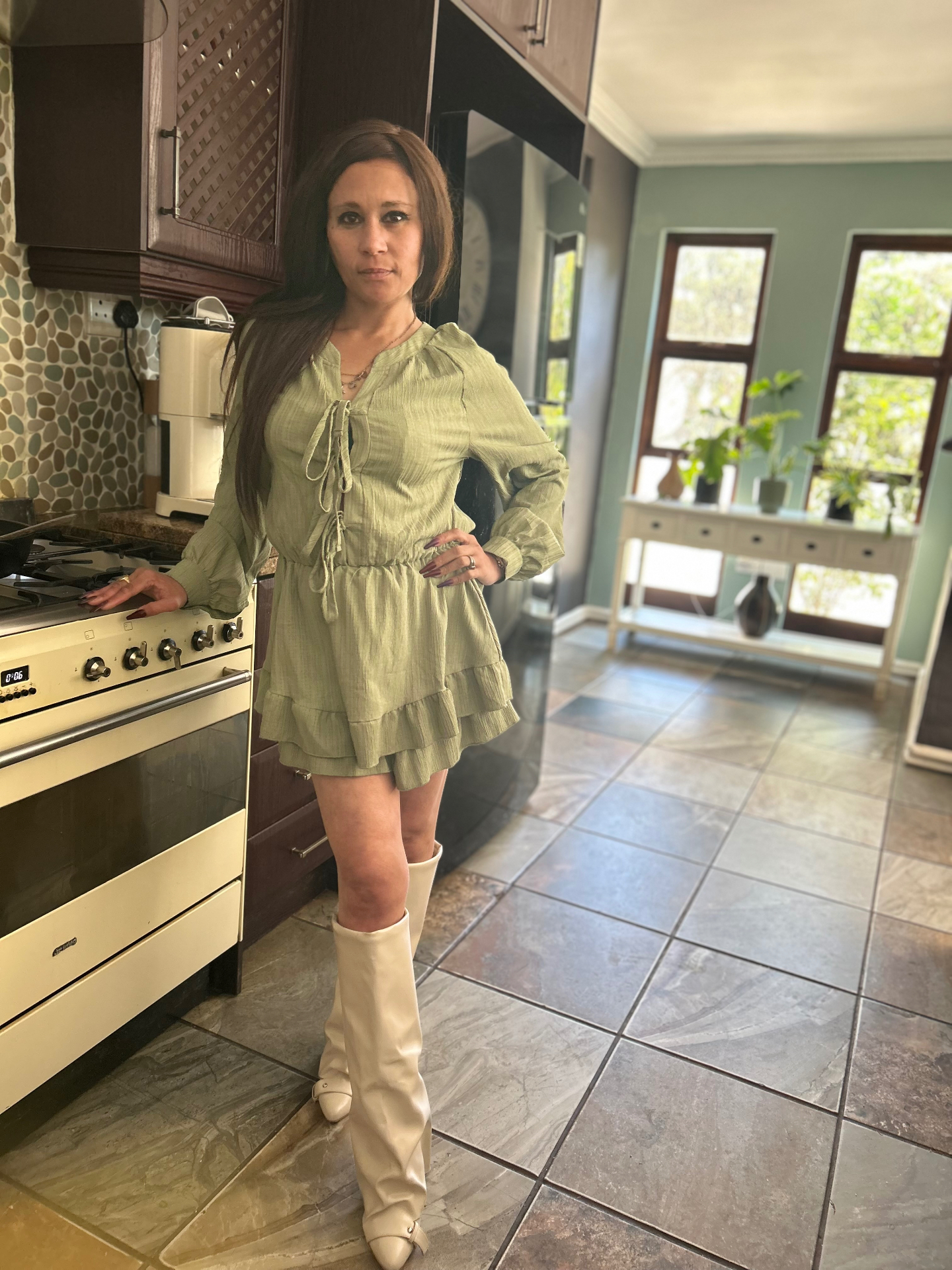 Cass in an olive-green tie-front ruffle playsuit and cream cone-heel knee boots, standing in her kitchen