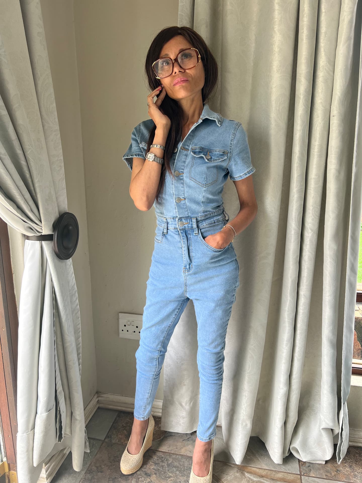 Cass in 2024 wearing a light-wash denim jumpsuit with oversized glasses and espadrille wedges