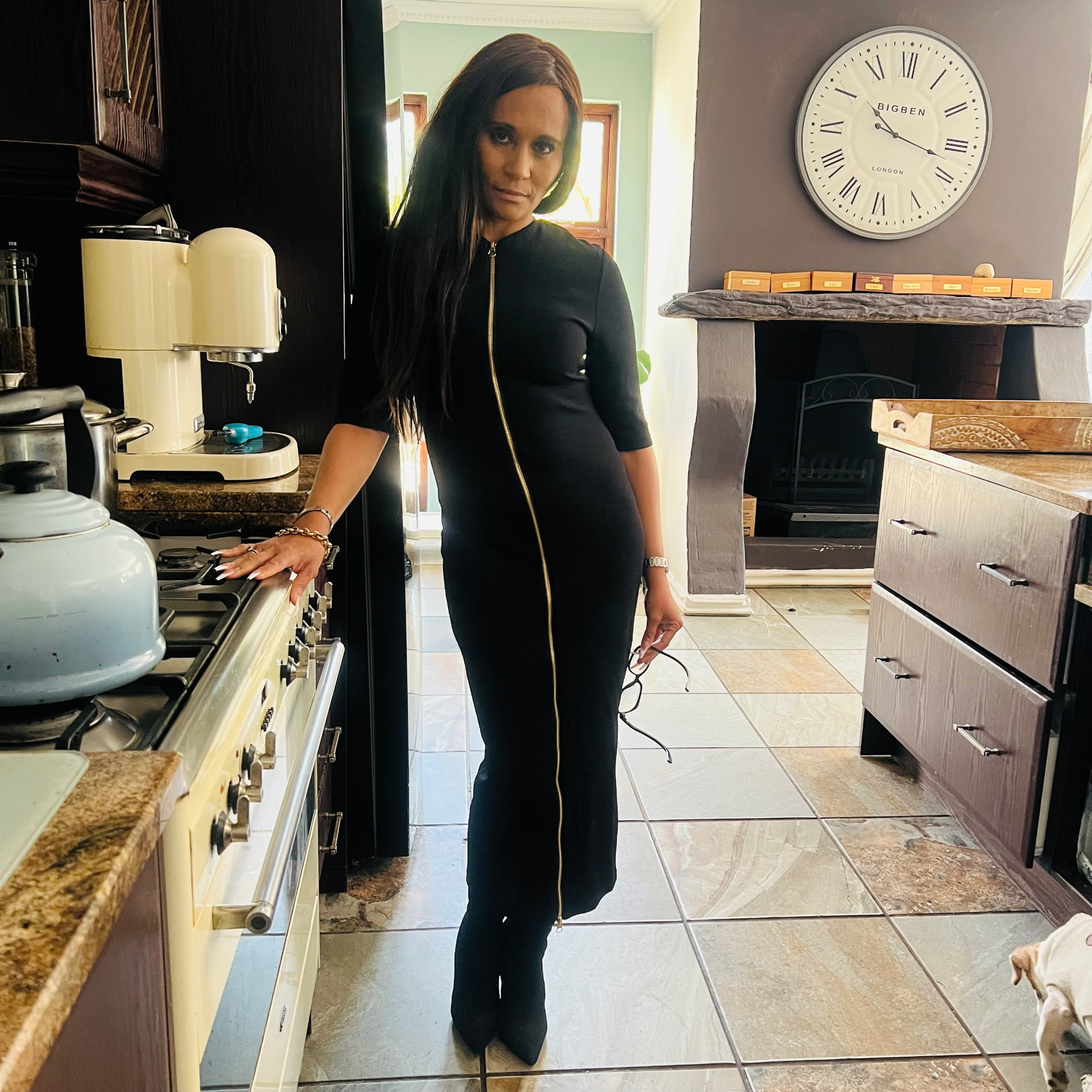 Cass in a long black fitted dress with gold double zips and black suede ankle boots, standing in her kitchen