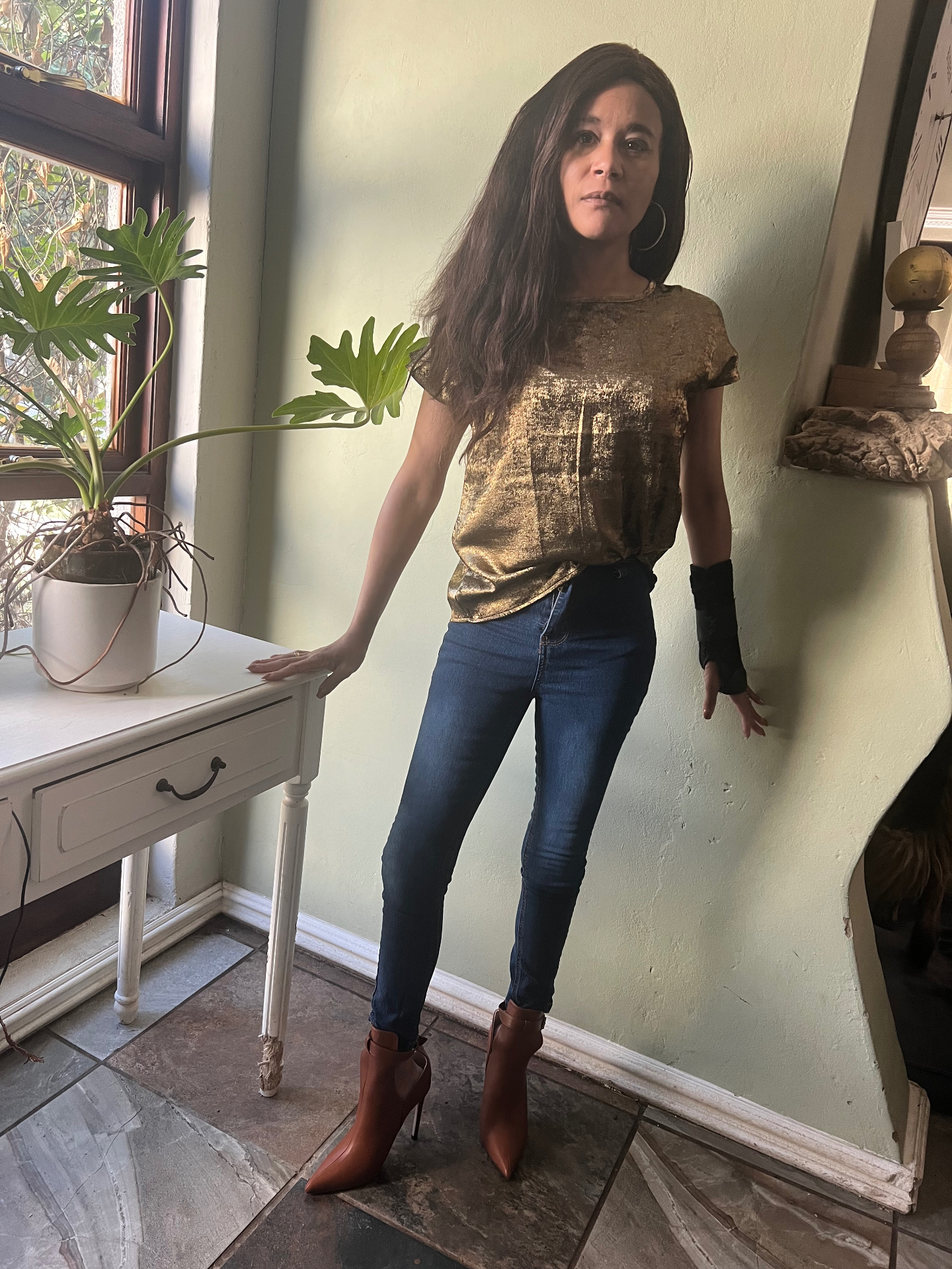 Cass in 2024, visibly underweight, wearing a gold sequin top, jeans and tan ankle boots, leaning against a white console table