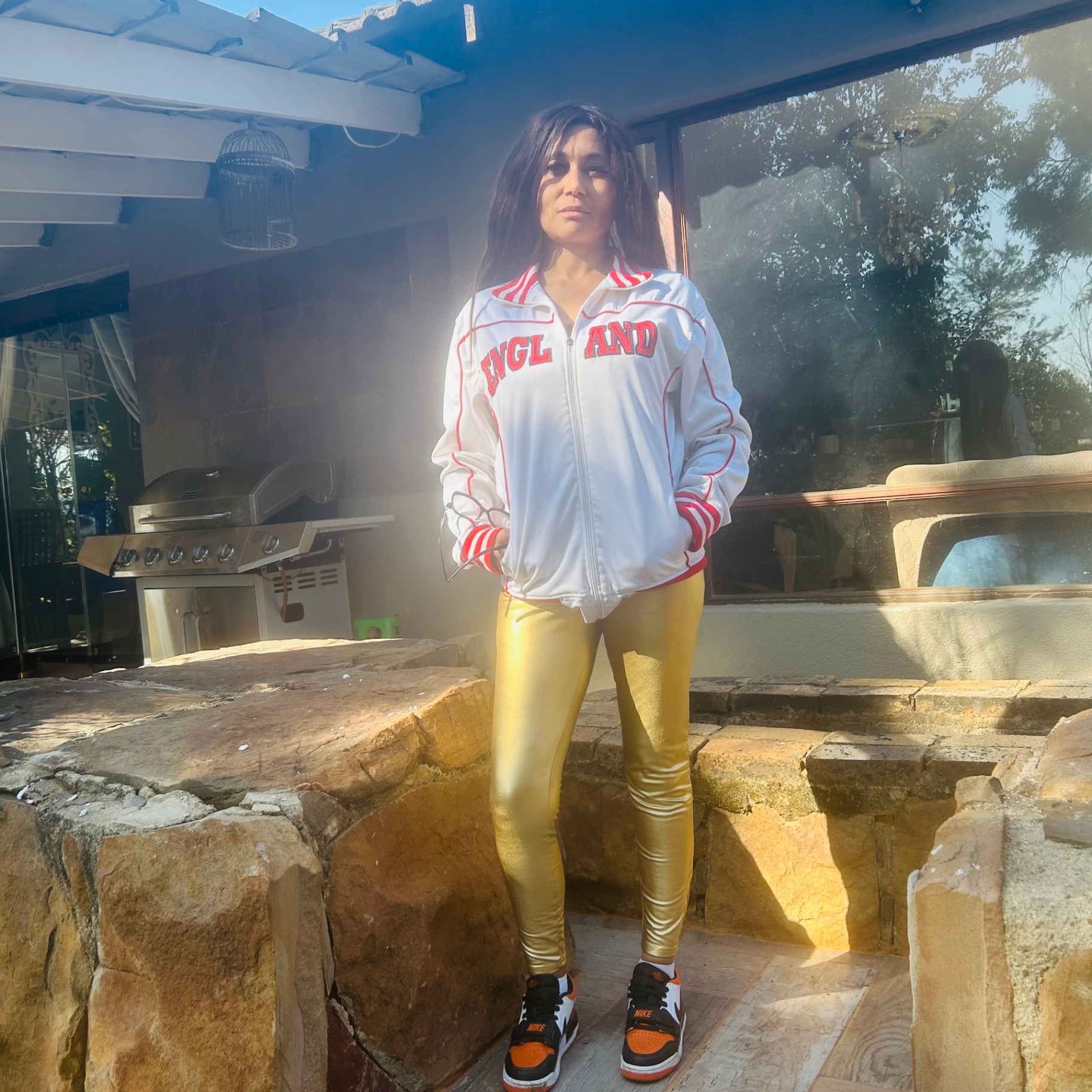 Cass in a white England track jacket, gold leggings and Nike high-tops, standing in a sunlit Jo'burg courtyard.