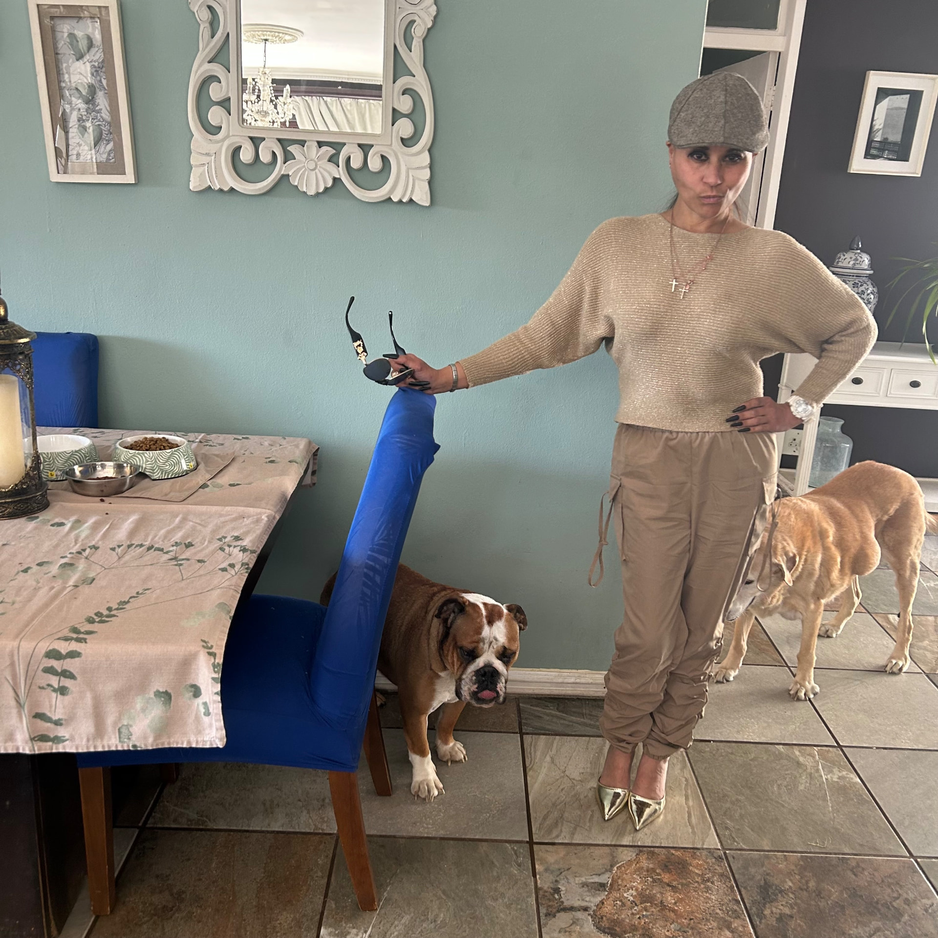 Cass in a gold-shimmer knit jumper, taupe cargo joggers, grey wool baker-boy cap and gold pointed heels, with a bulldog and a labrador beside her