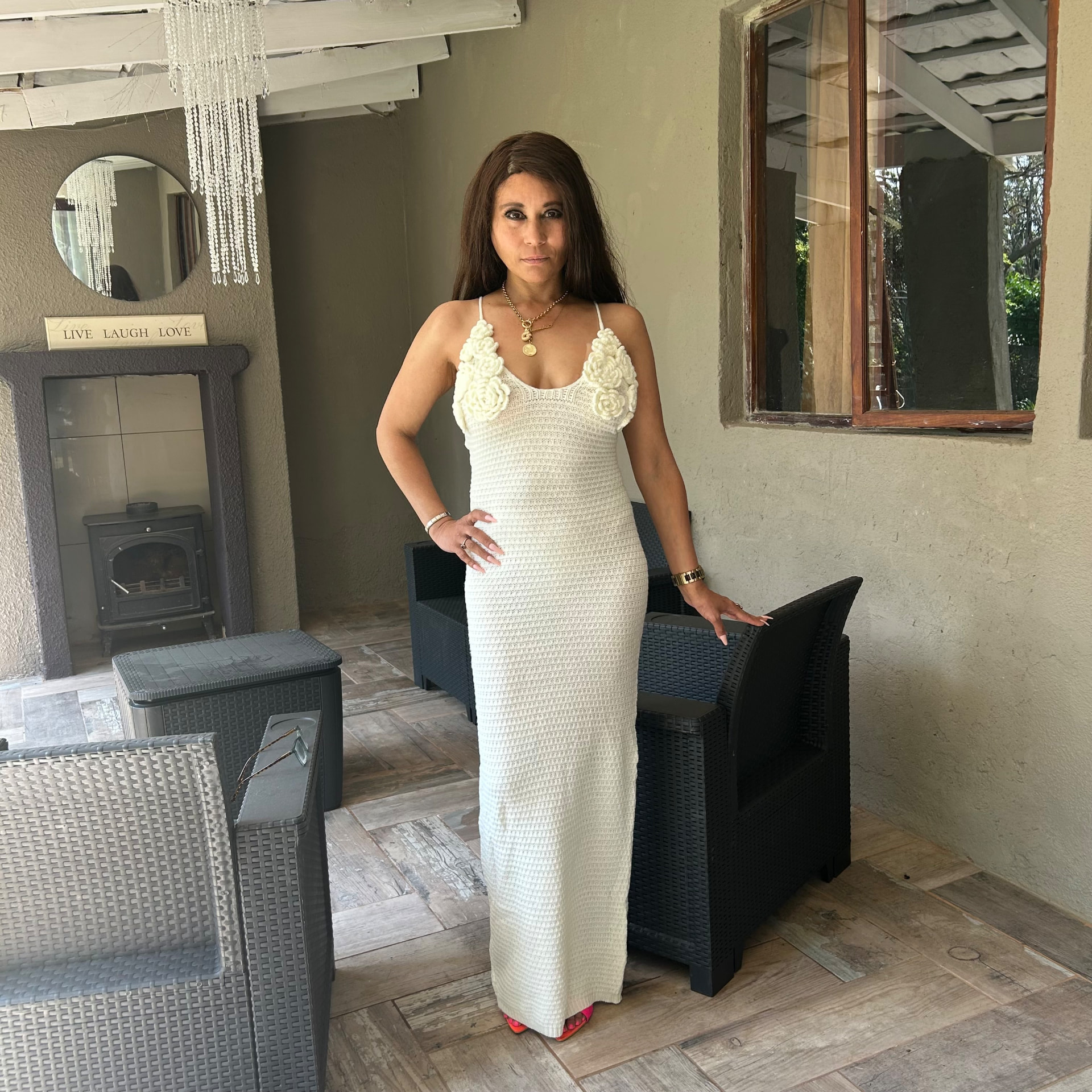 Cass in an ivory crochet maxi dress with rose appliqués on the straps, pink sandals and gold jewellery, on the patio