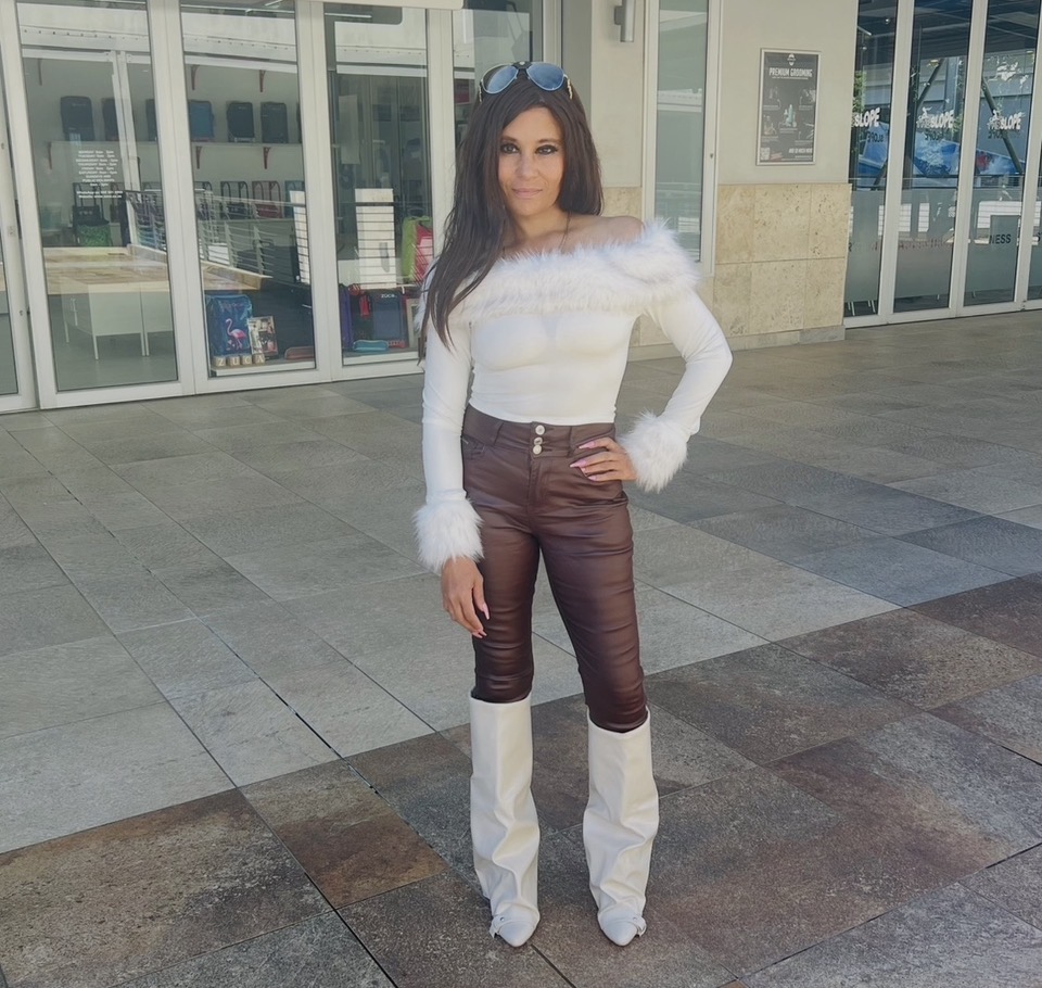 Cass standing tall in a white off-shoulder fur-trim top, chocolate leather trousers and slouched white knee-high boots, sunglasses on her head