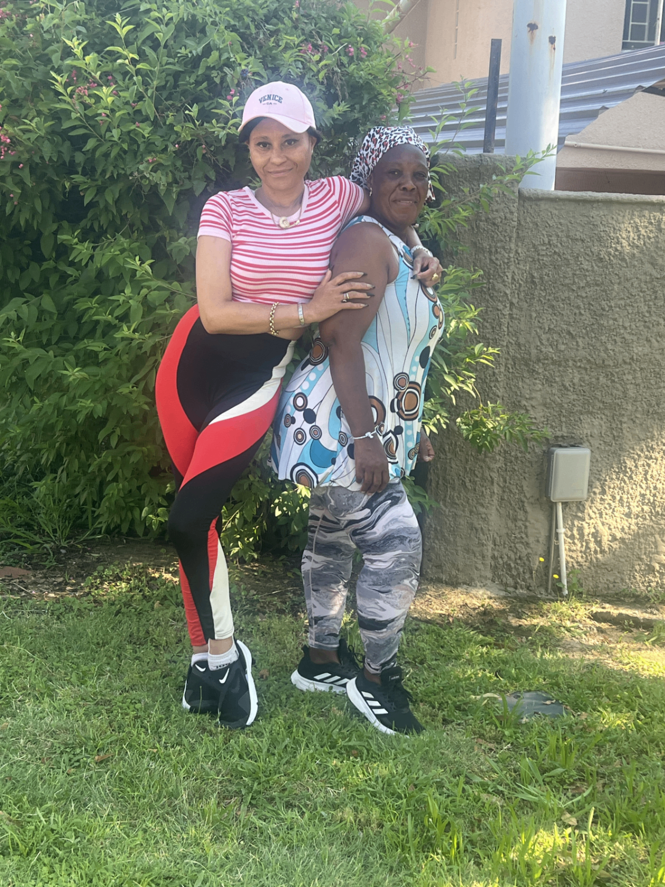 Cass in a pink Venice cap and striped tee, leg lifted in a playful stretch pose, arm around her helper Ntombi in a colourful print top — the two of them in a sunlit garden