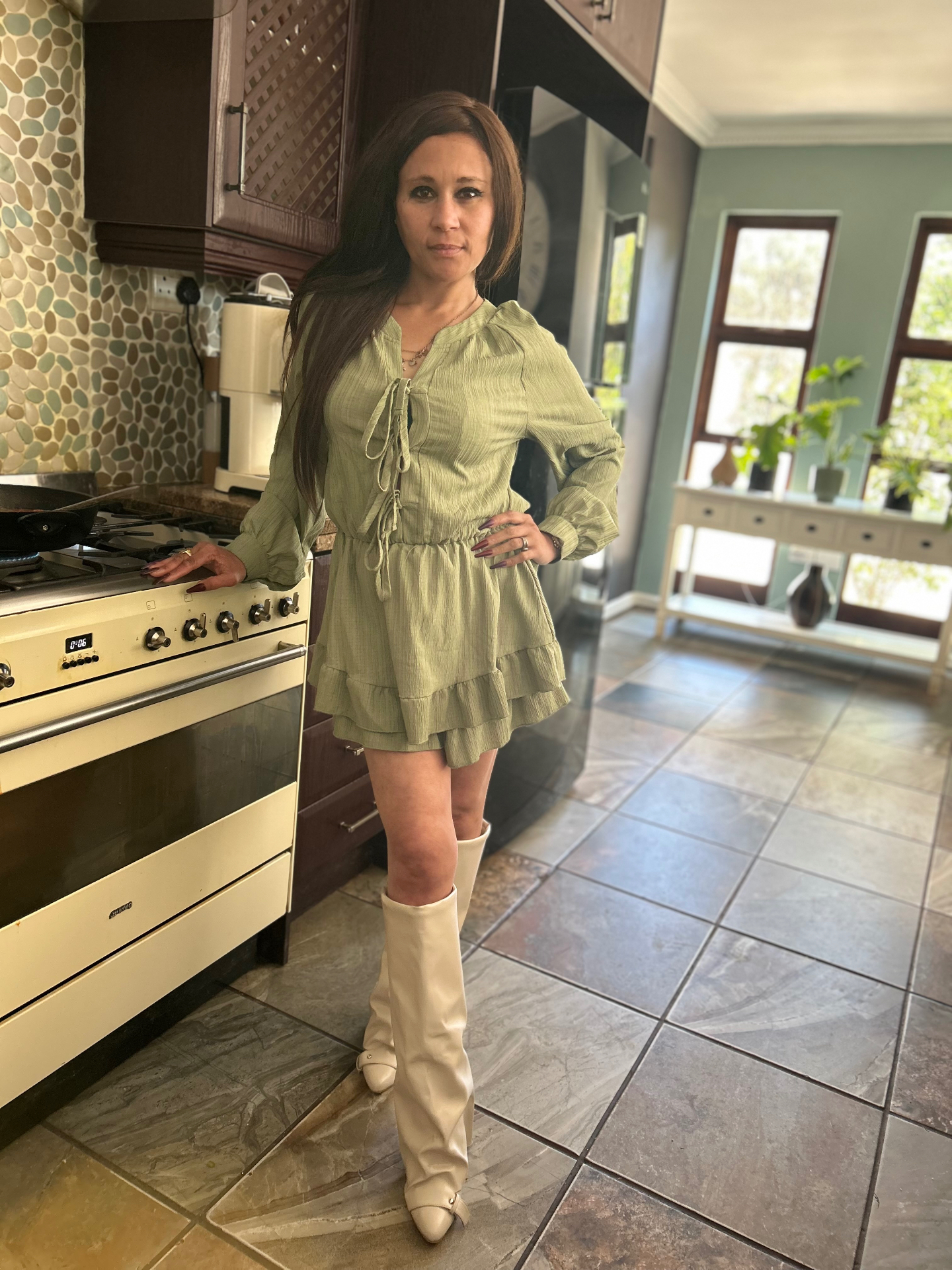 Cass in an olive-green tie-front ruffle playsuit and cream cone-heel knee boots, standing in her kitchen