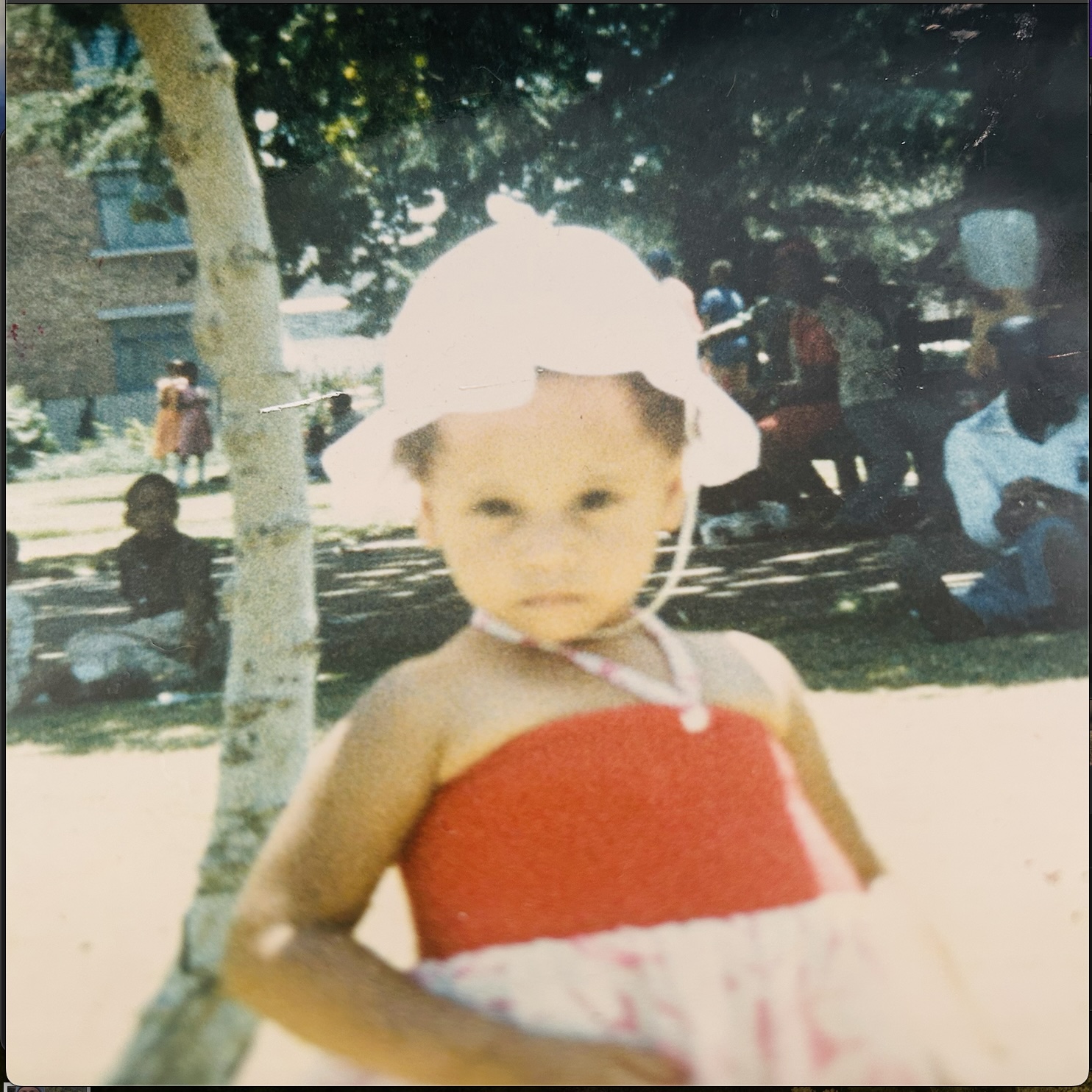 Cass, aged four, in a white sunhat and red sundress, staring straight down the barrel of the camera.