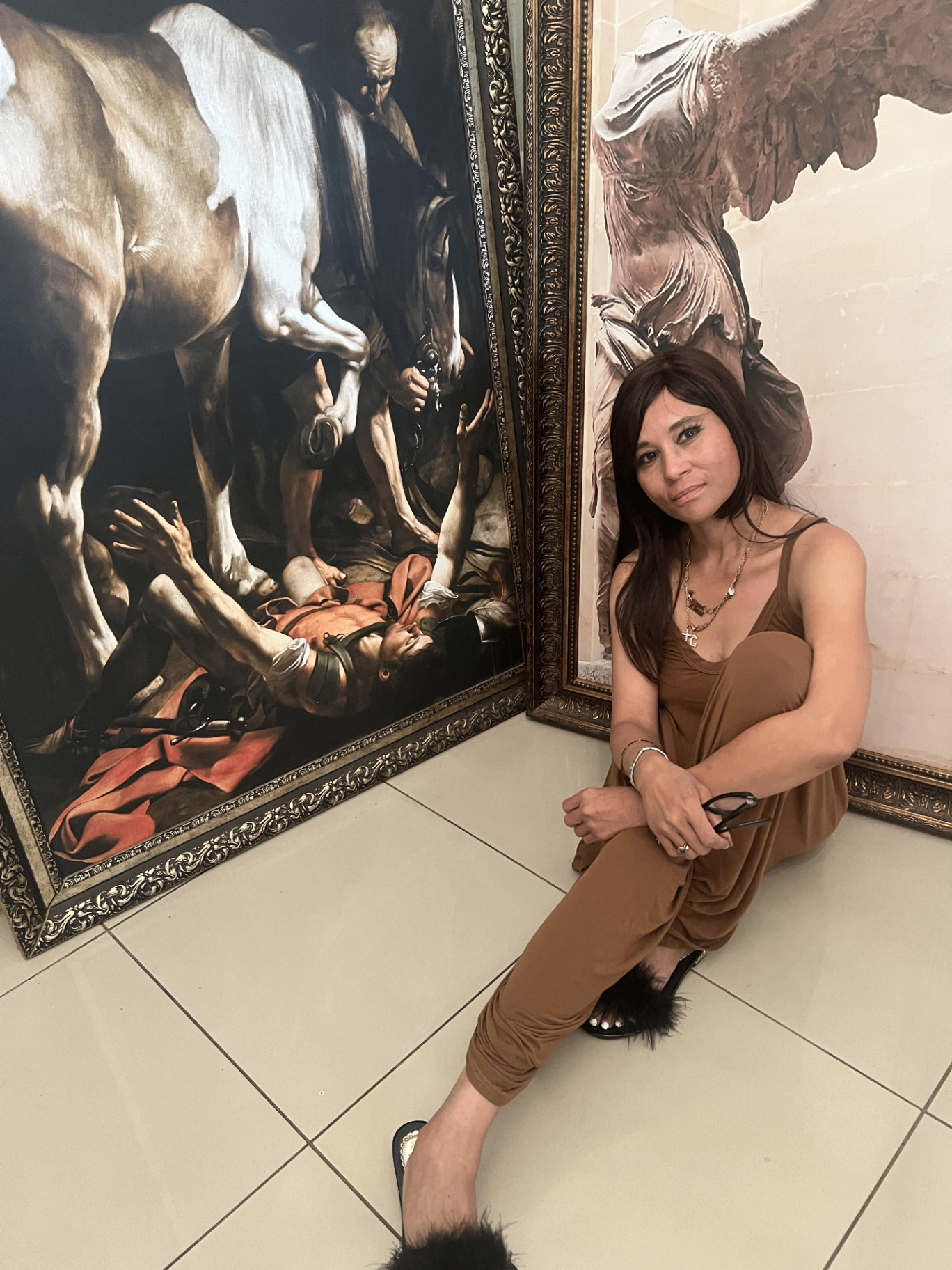 Cass sitting on a tiled floor in a caramel jumpsuit and black feather slides, framed between a large print of Caravaggio's Conversion of Saint Paul and a print of the Winged Victory of Samothrace (Nike).