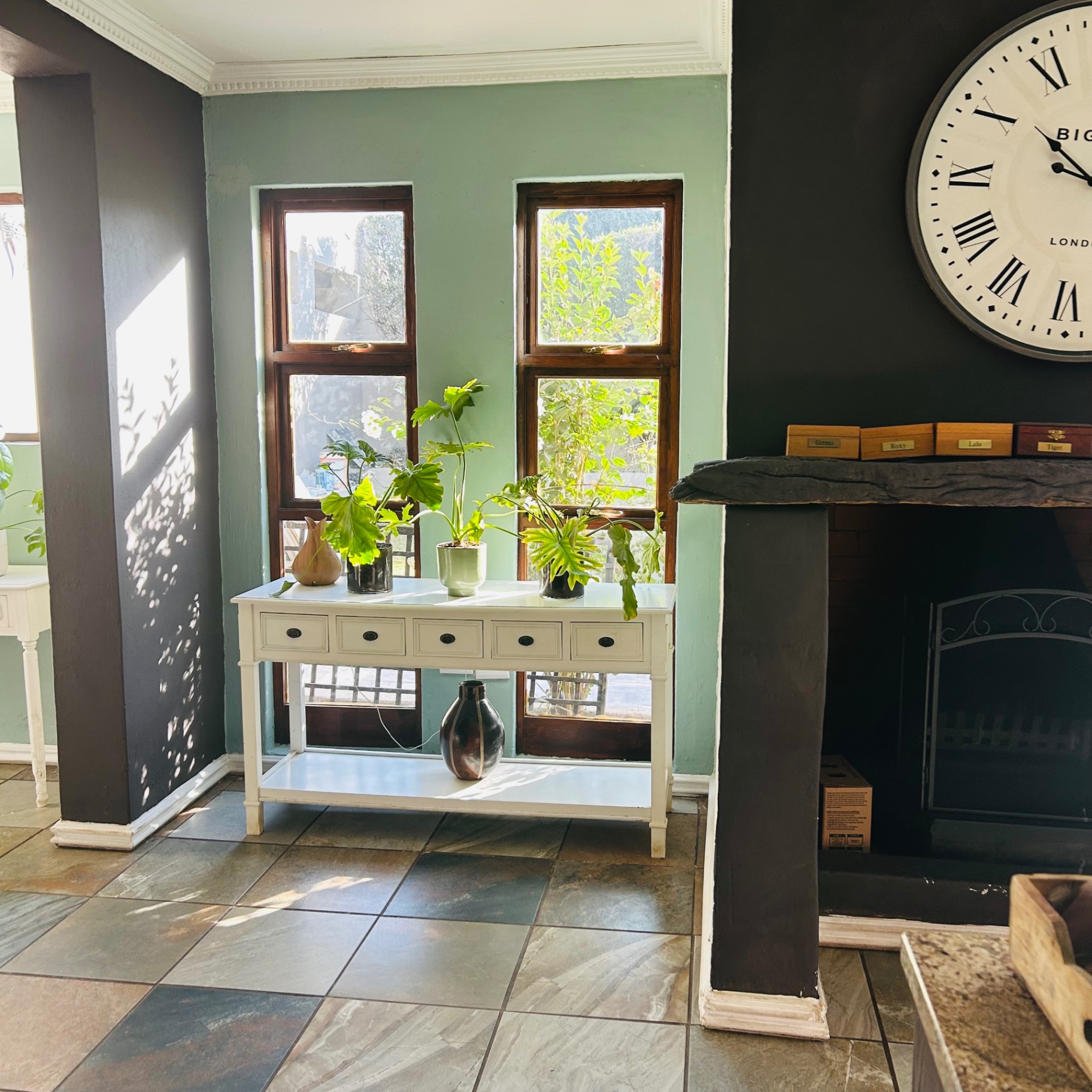 Sage green wall with tall windows, white console of plants and inky fireplace with Big Ben clock