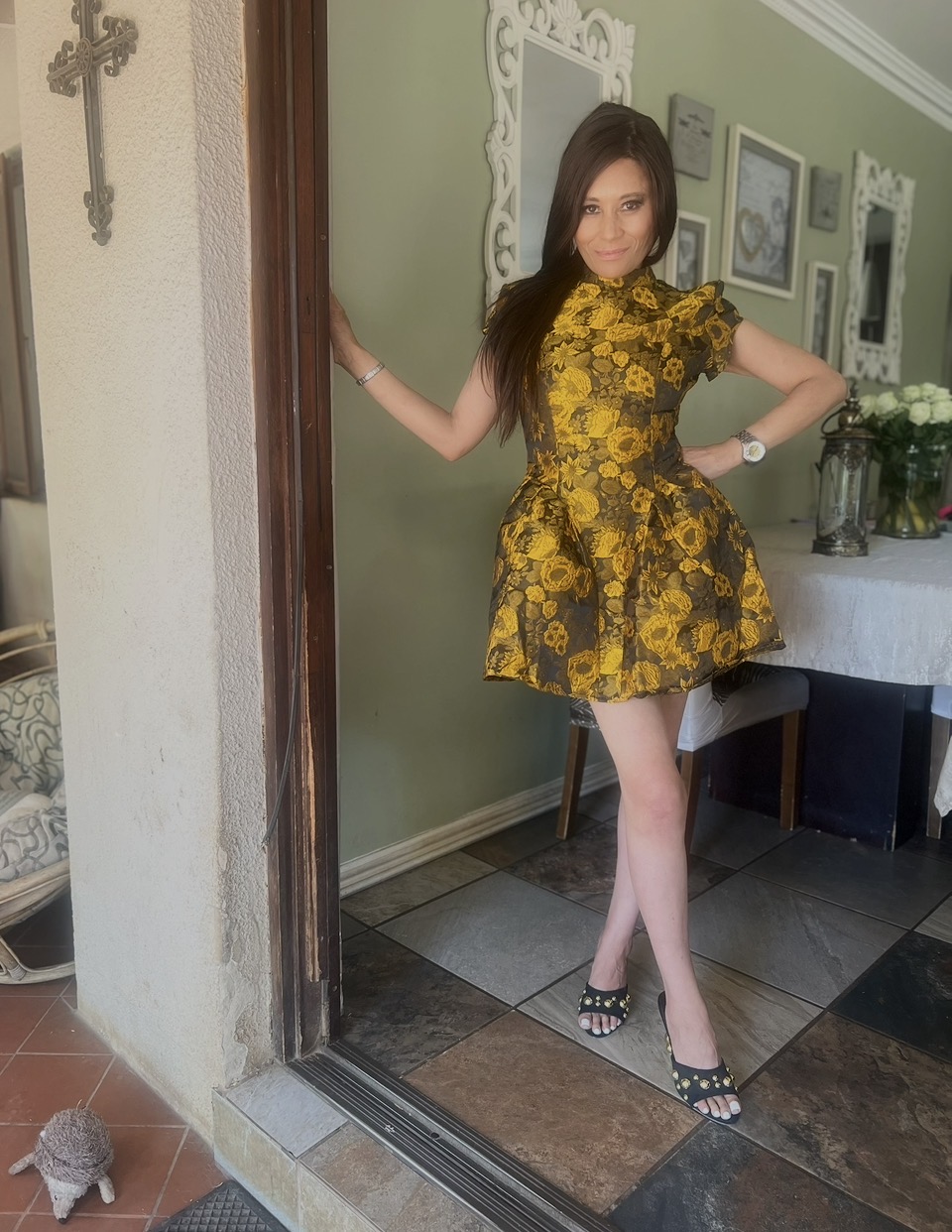 Cass in a gold and black brocade mini dress with studded slides, smiling in the doorway
