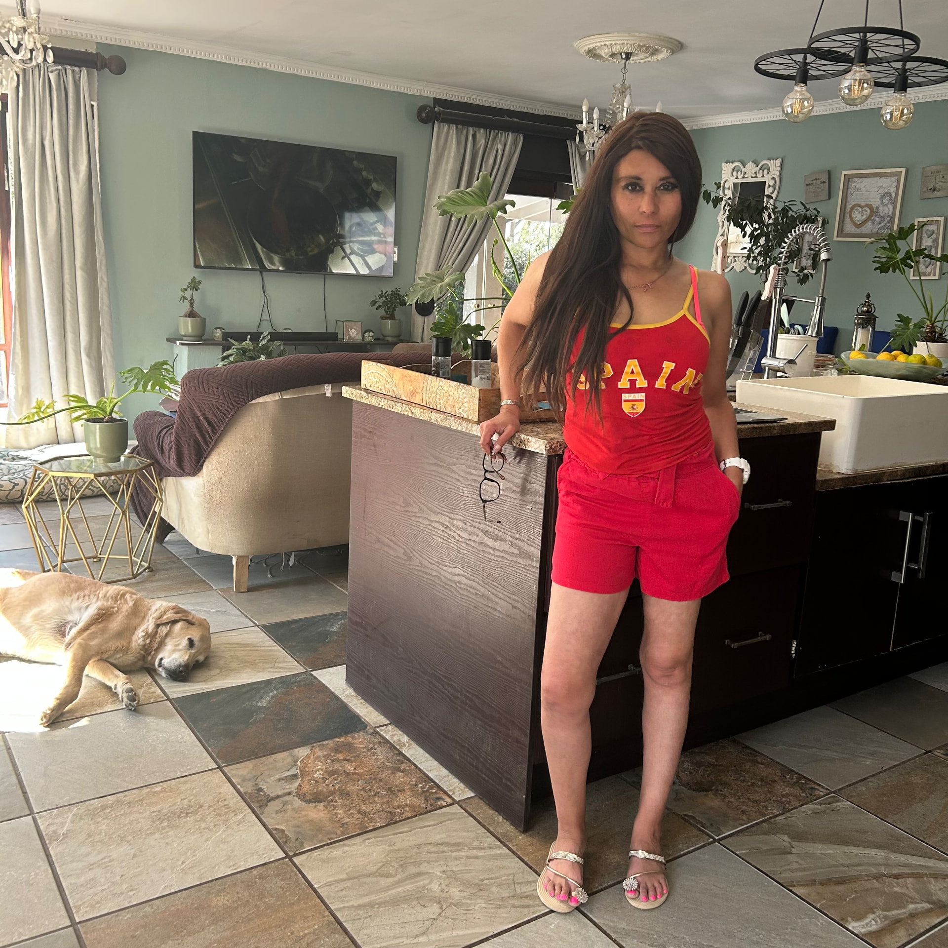 Cass in a red Spain tank top and shorts with silver embellished sandals, a golden retriever asleep on the tiled floor