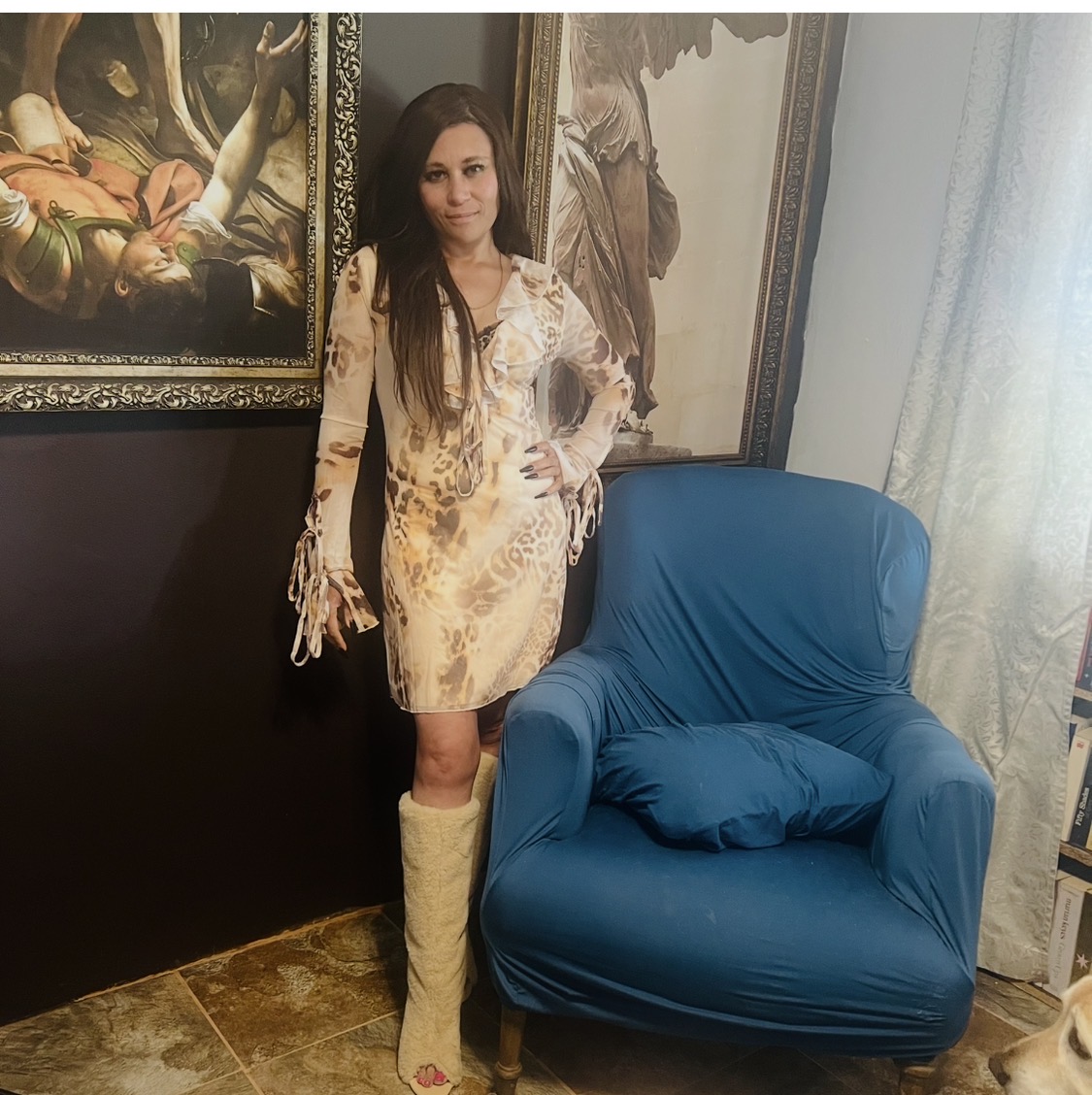 Cass in a ruffled leopard chiffon mini dress and cream peep-toe furry boots, standing between two large framed old-master paintings beside a teal armchair