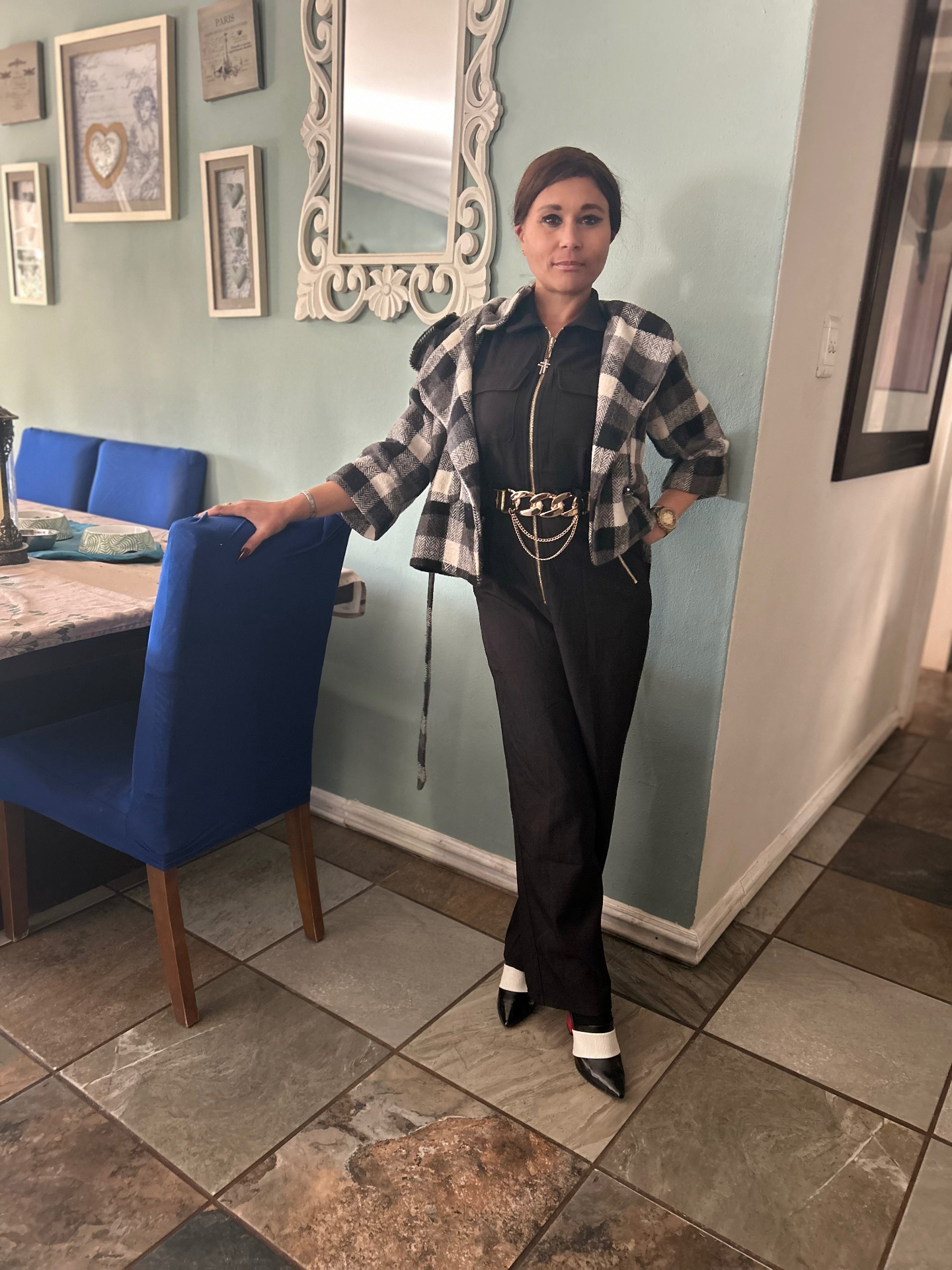Cass in the black jumpsuit and checkered jacket standing beside a blue dining chair