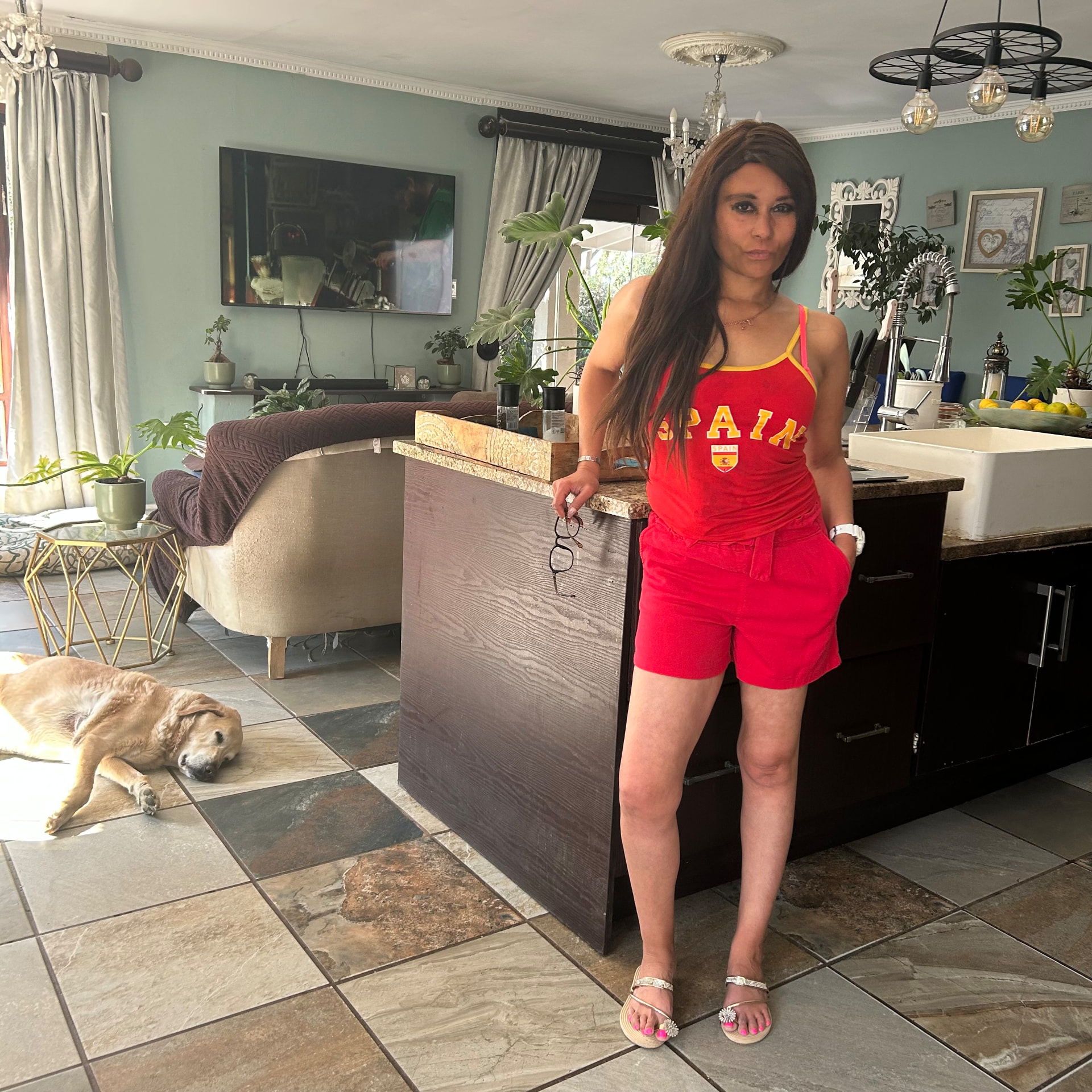 Cass in a red Spain tank top and matching red shorts, holding her glasses, standing in the kitchen