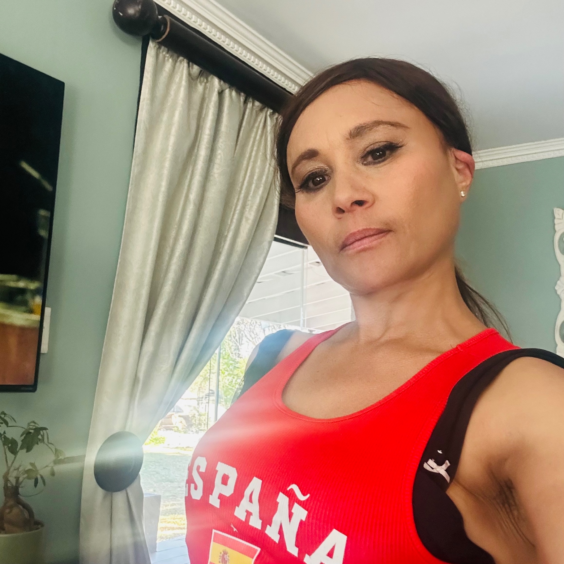Cass in a red España vest top, hair back, soft Sunday makeup, sage-green wall behind her
