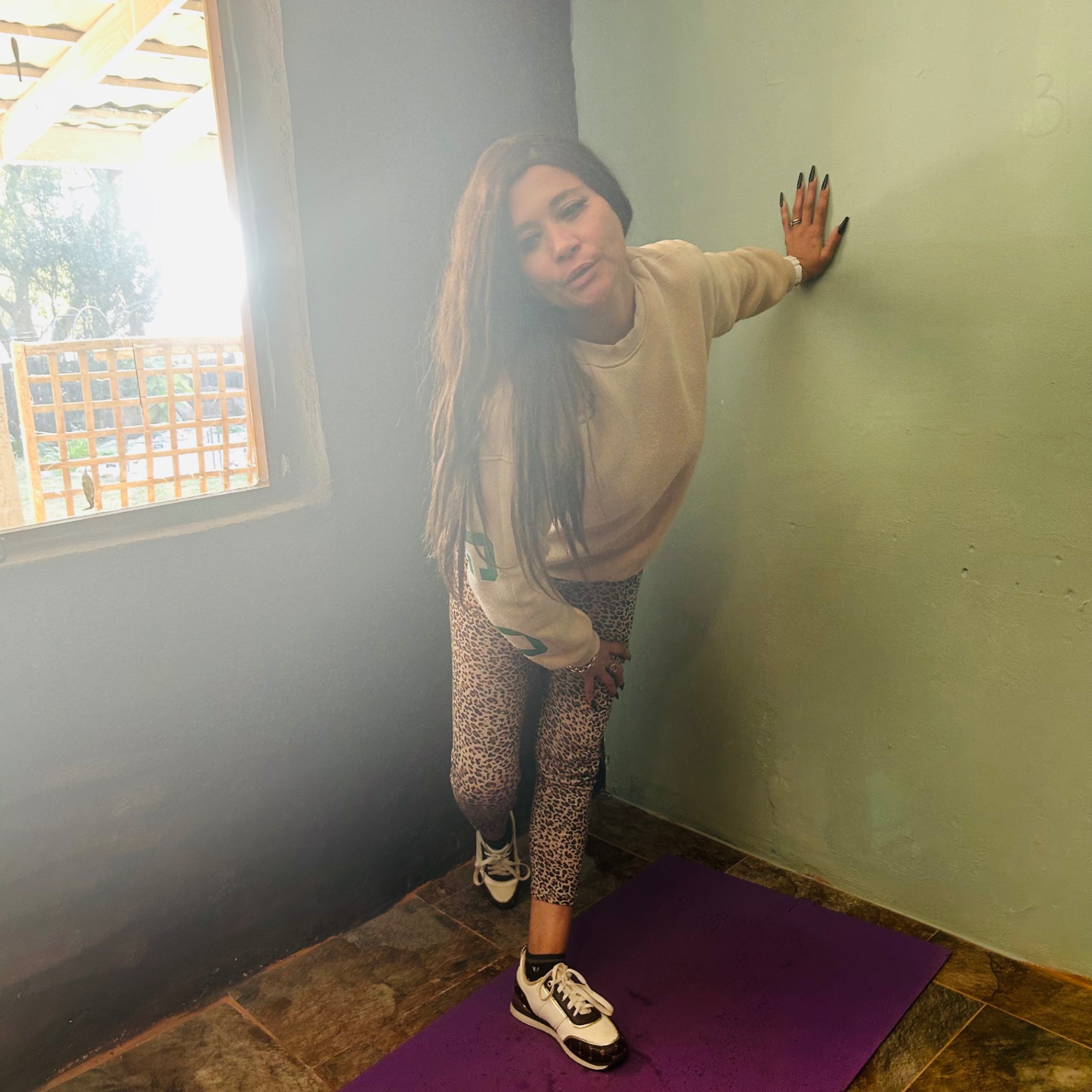 Cass in a cream sweatshirt and leopard-print leggings, leaning against a sage wall in an unfinished room, mid tai chi.