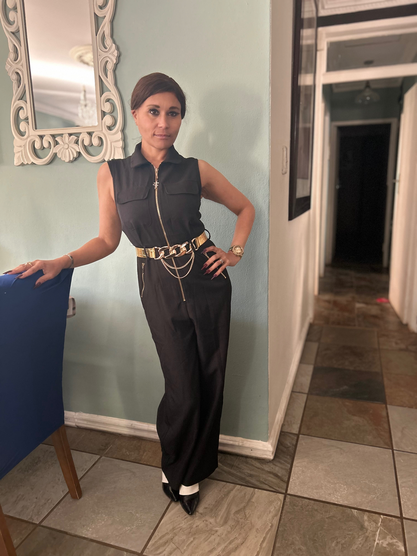 Cass in a sleeveless zip-front black jumpsuit with a gold chain belt and black pointed heels with white ankle cuffs, standing against a sage-green wall