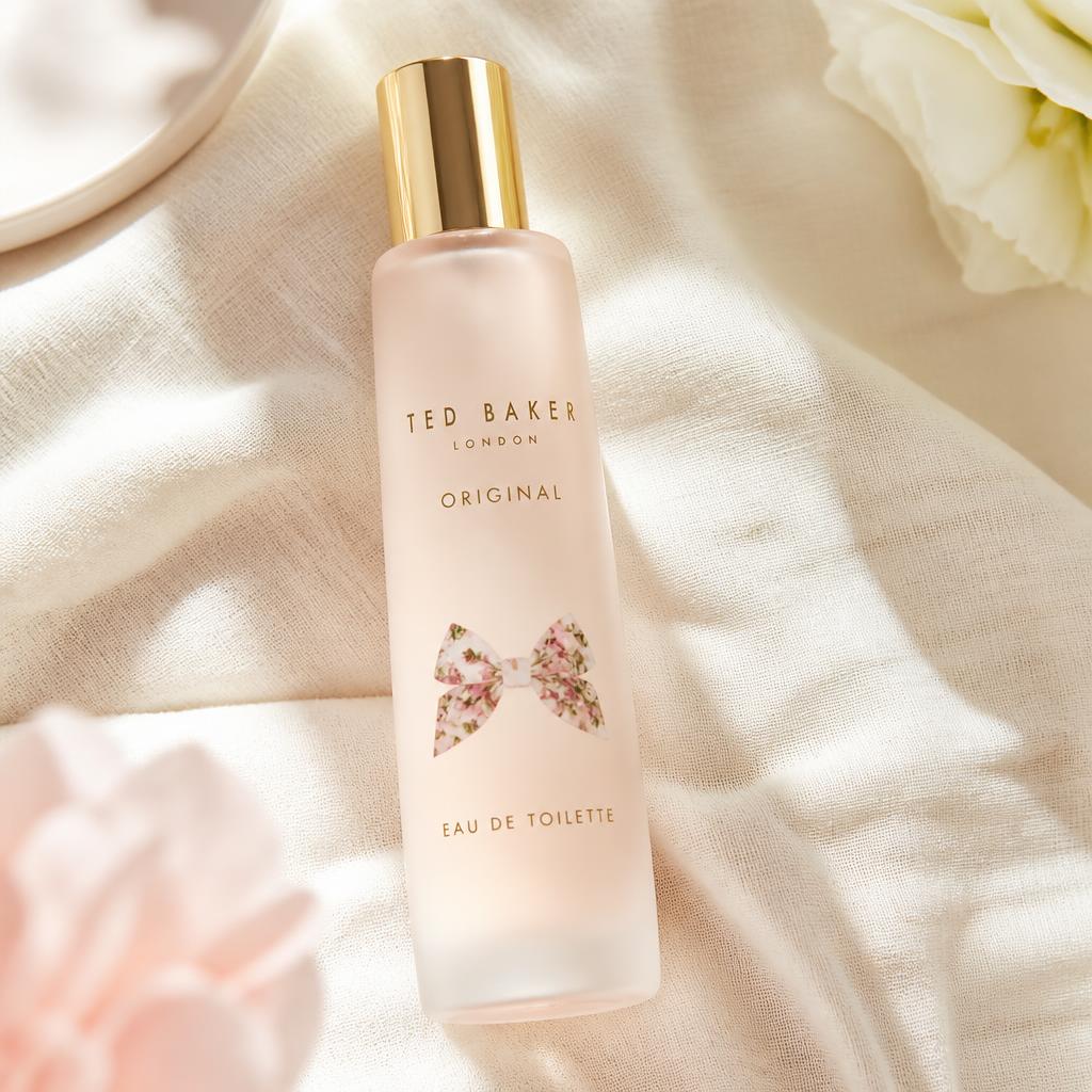 Ted Baker Original eau de toilette bottle in frosted blush glass with a gold cap and floral bow, on cream linen